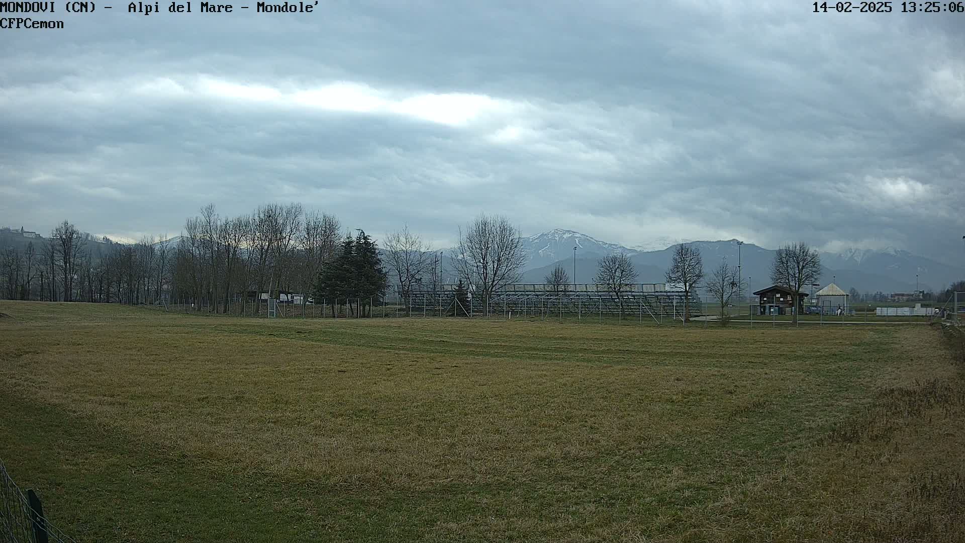 A field of brown-green grass stretches toward bleachers and a small building, with bare trees and snow-capped mountains under a cloudy sky in the background.