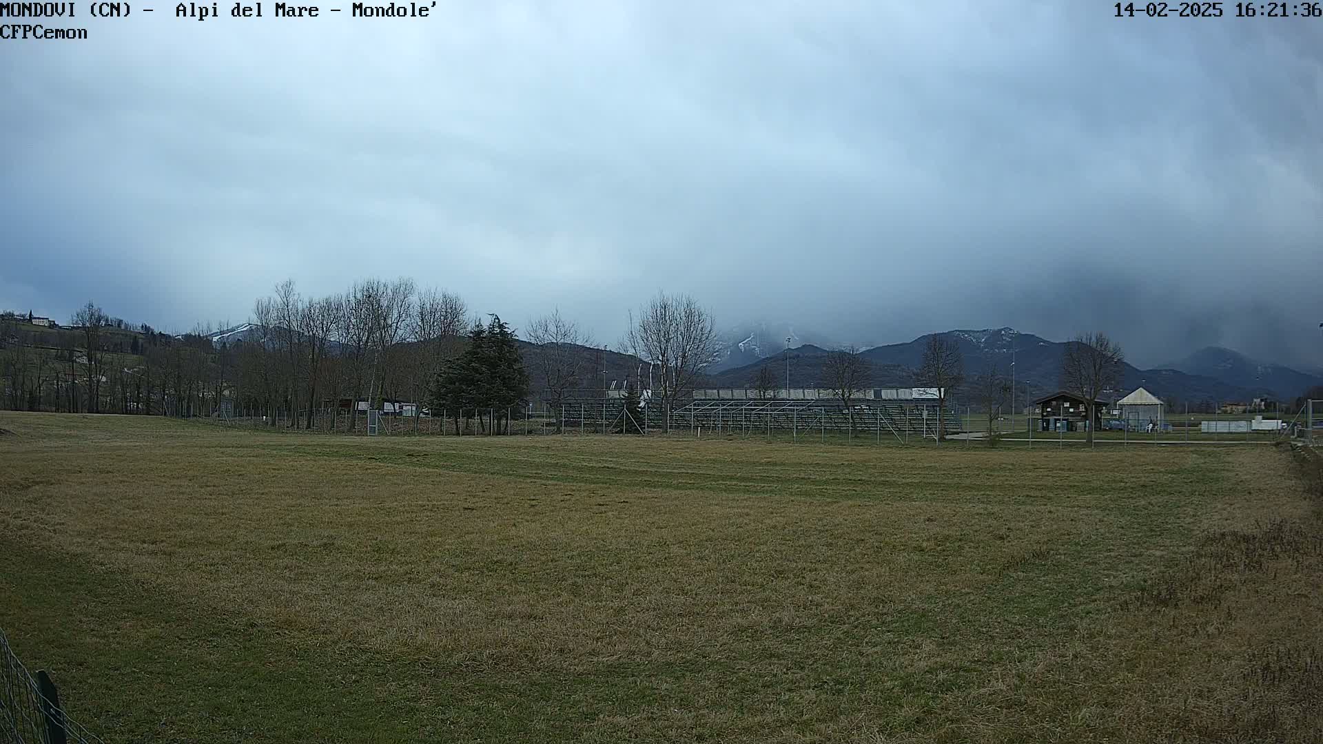 A mostly cloudy day shows a grassy field with bleachers and a small building in the background, beyond which are snow-capped mountains.