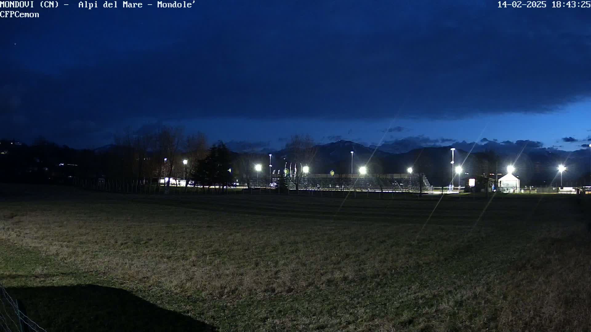 A dark-blue twilight sky hangs over a field, beyond which are illuminated bleachers and a small building, all set against a backdrop of distant, snow-capped mountains.