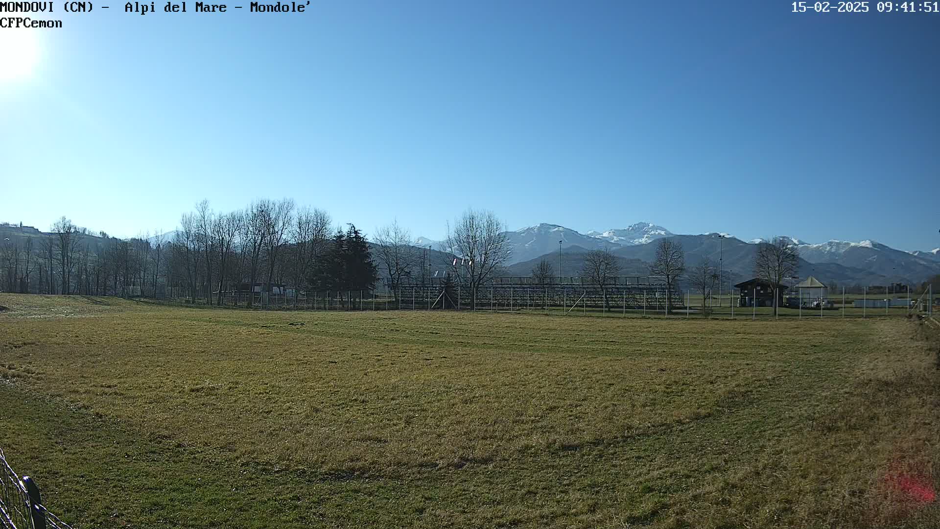 A grassy field with bleachers and a small building sits in the foreground, with snow-capped mountains visible in the background under a clear blue sky.
