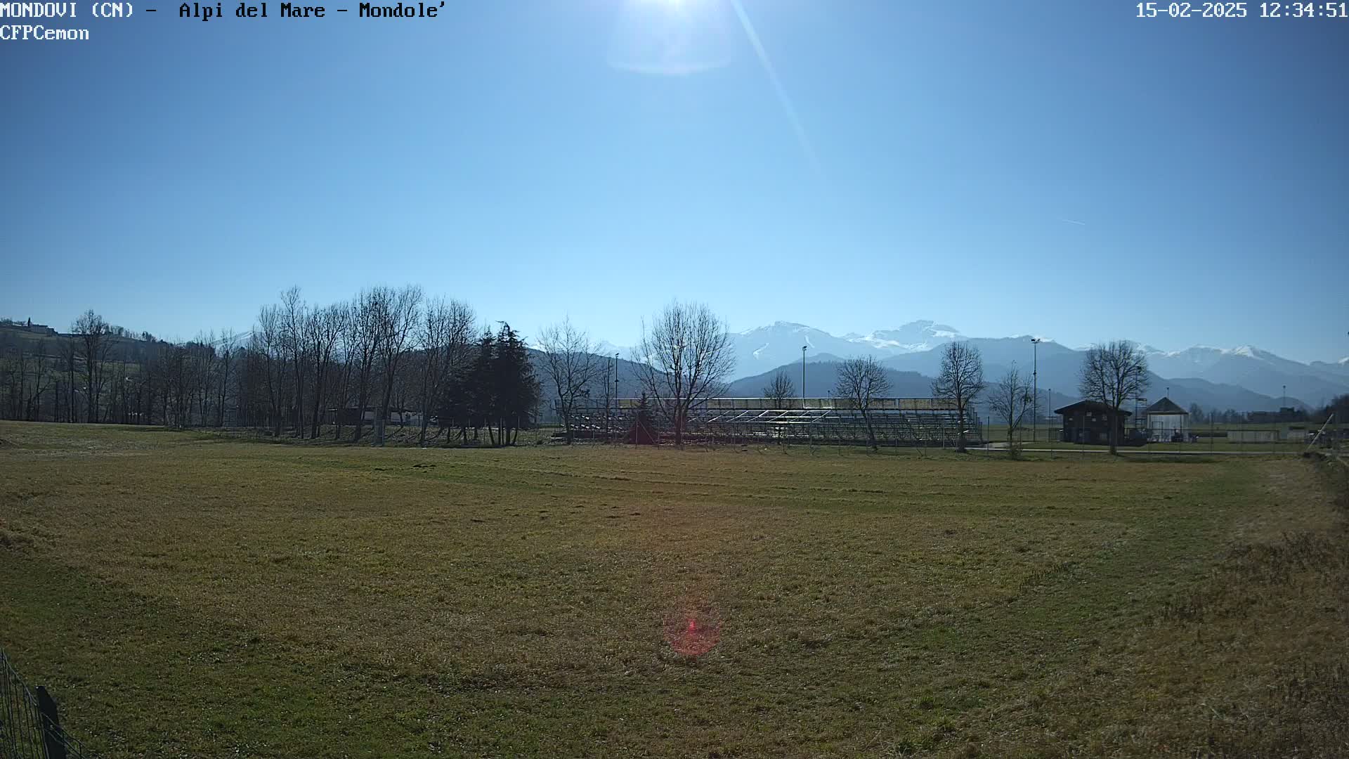 A sunny day reveals a grassy field with leafless trees, a small building, and bleachers in the foreground, all set against a backdrop of snow-capped mountains under a clear blue sky.