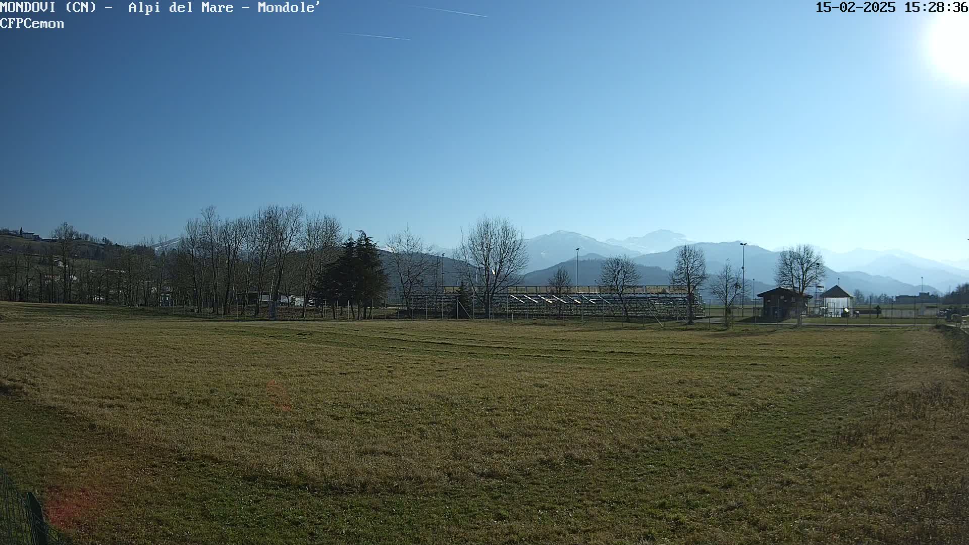 A grassy field with a small building and empty bleachers sits in the foreground, under a clear blue sky, with mountains visible in the background.