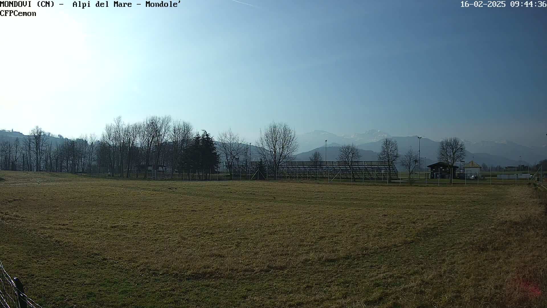 A brown grassy field stretches out before empty bleachers and a small building, with bare trees and snow-capped mountains visible in the distance under a clear blue sky.