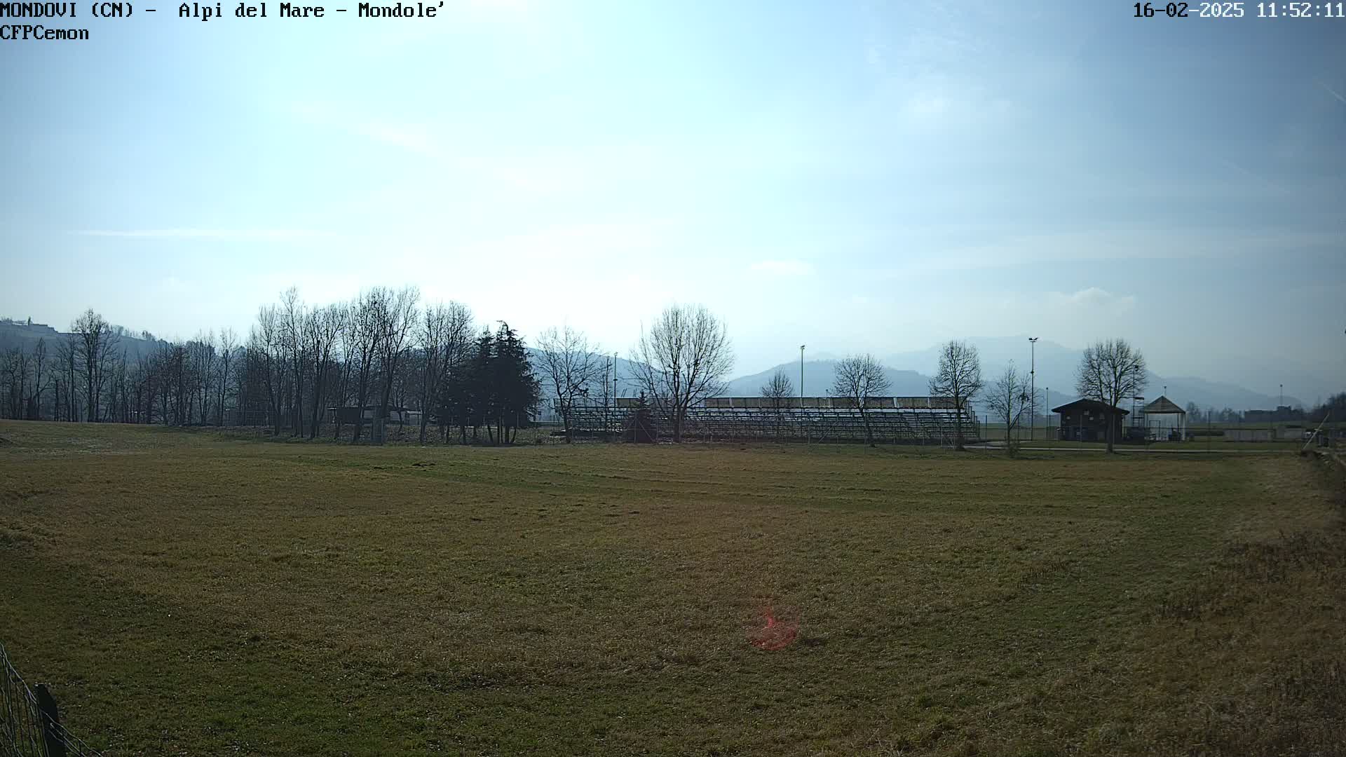 A mostly sunny day reveals a brown grassy field, leafless trees, a small building, and bleachers against a backdrop of distant, hazy mountains.