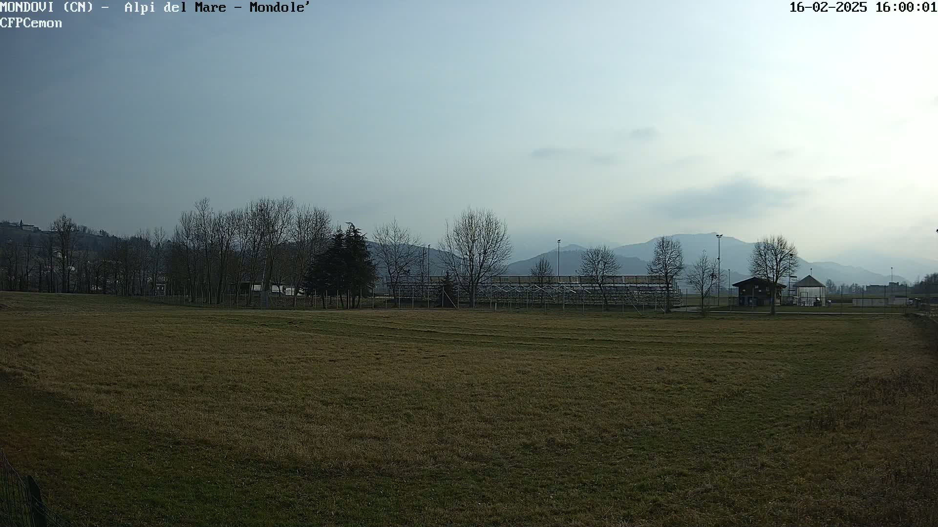 A brown, grassy field stretches towards bleachers and a small building, backed by bare trees and hazy mountains under a pale sky.