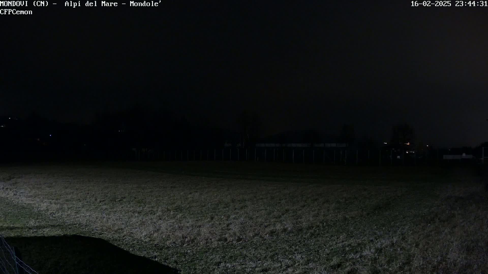 A dark, nighttime view of a grassy field enclosed by a fence, with dimly lit structures and trees visible in the distance under a cloudy sky.
