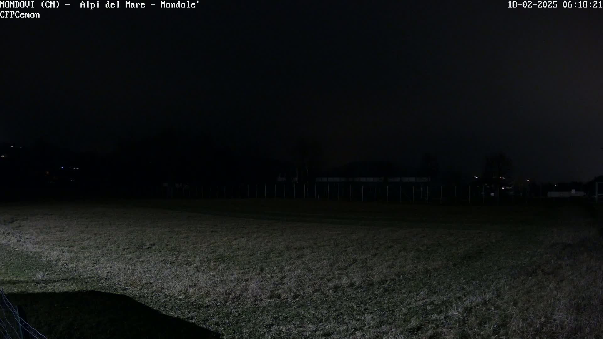 A dark, nighttime view of a grassy field, enclosed by a fence, with a few distant lights visible under a mostly clear, dark sky.
