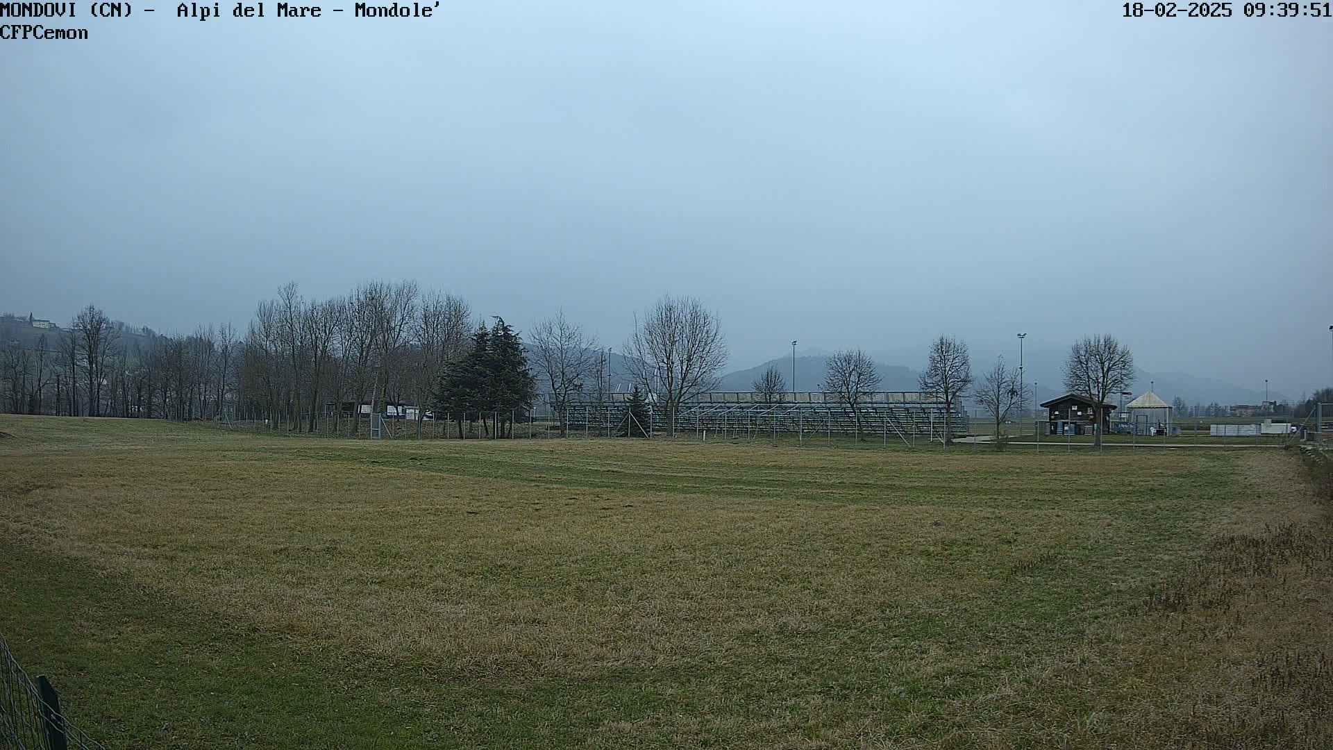 A mostly-brown field stretches out before bleachers and a small building, all set against a background of bare trees and hazy mountains under an overcast sky.