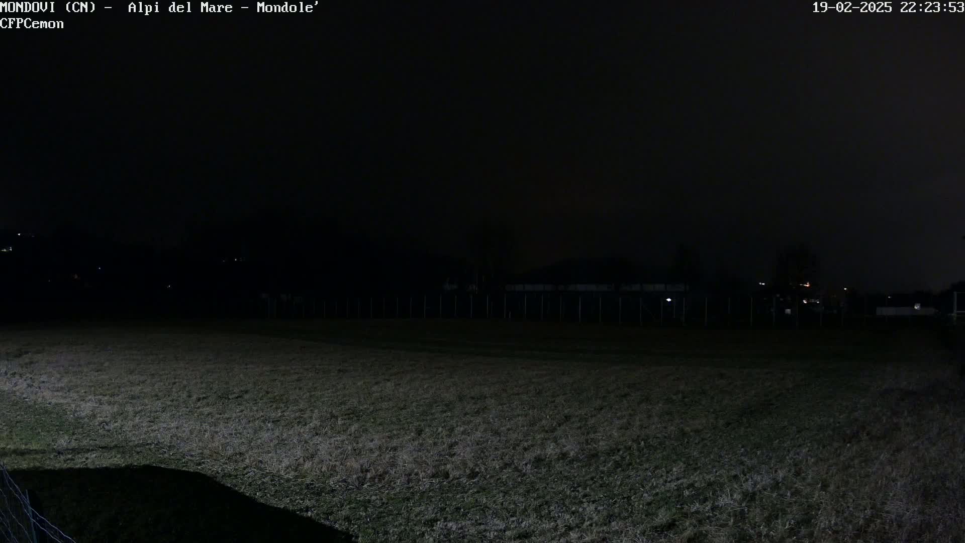 A dark, nighttime view of a grassy field enclosed by a fence, with a few distant lights faintly visible in the background.