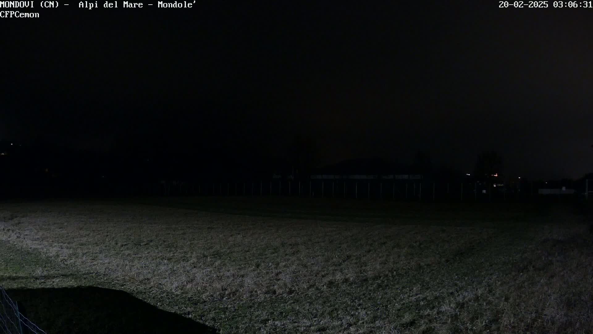 A dark, nighttime view of a grassy field with a fence and dimly lit structures in the distance.
