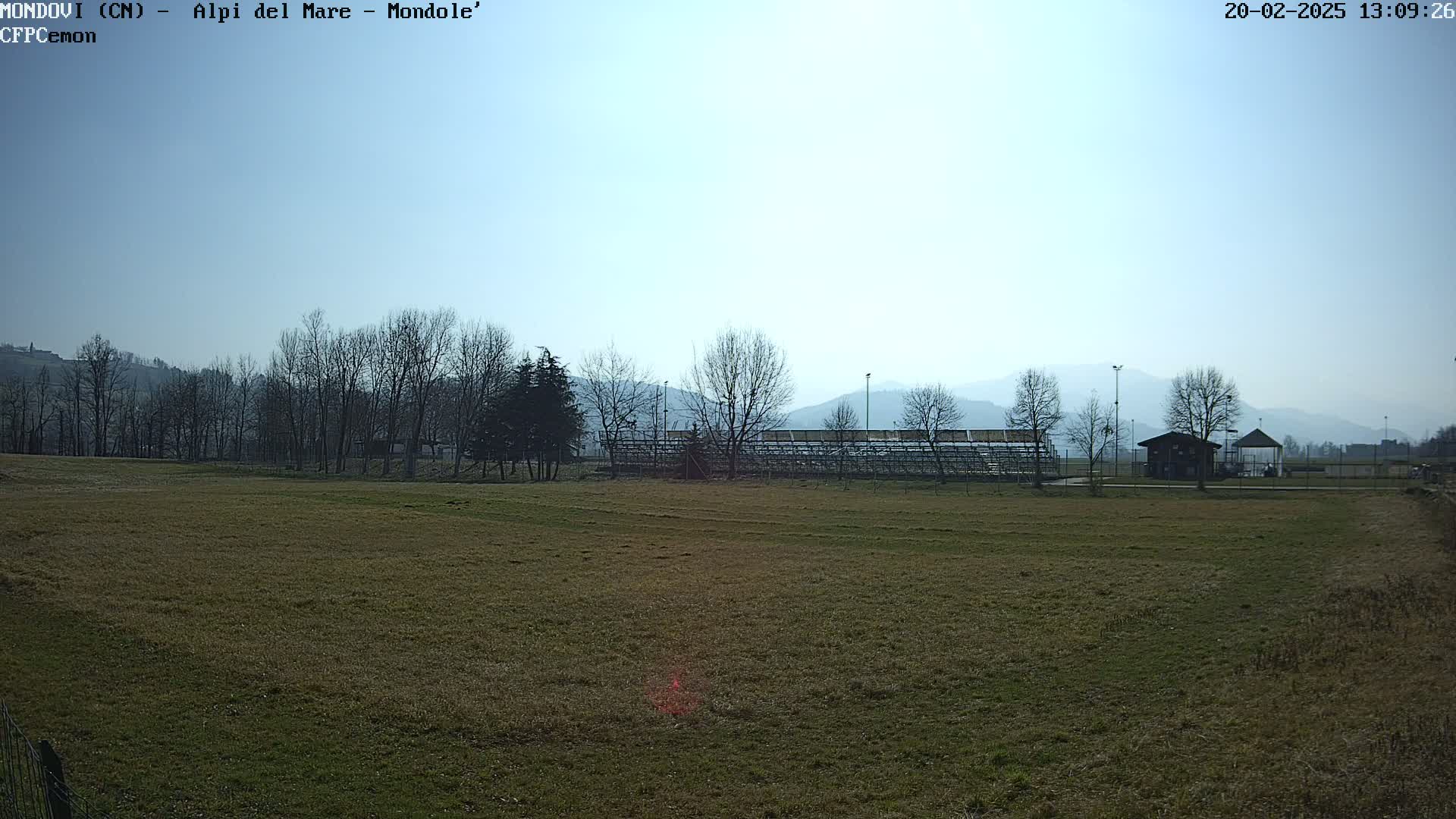 A grassy field stretches toward bleachers and a small building, backed by bare trees and hazy mountains under a clear, bright sky.