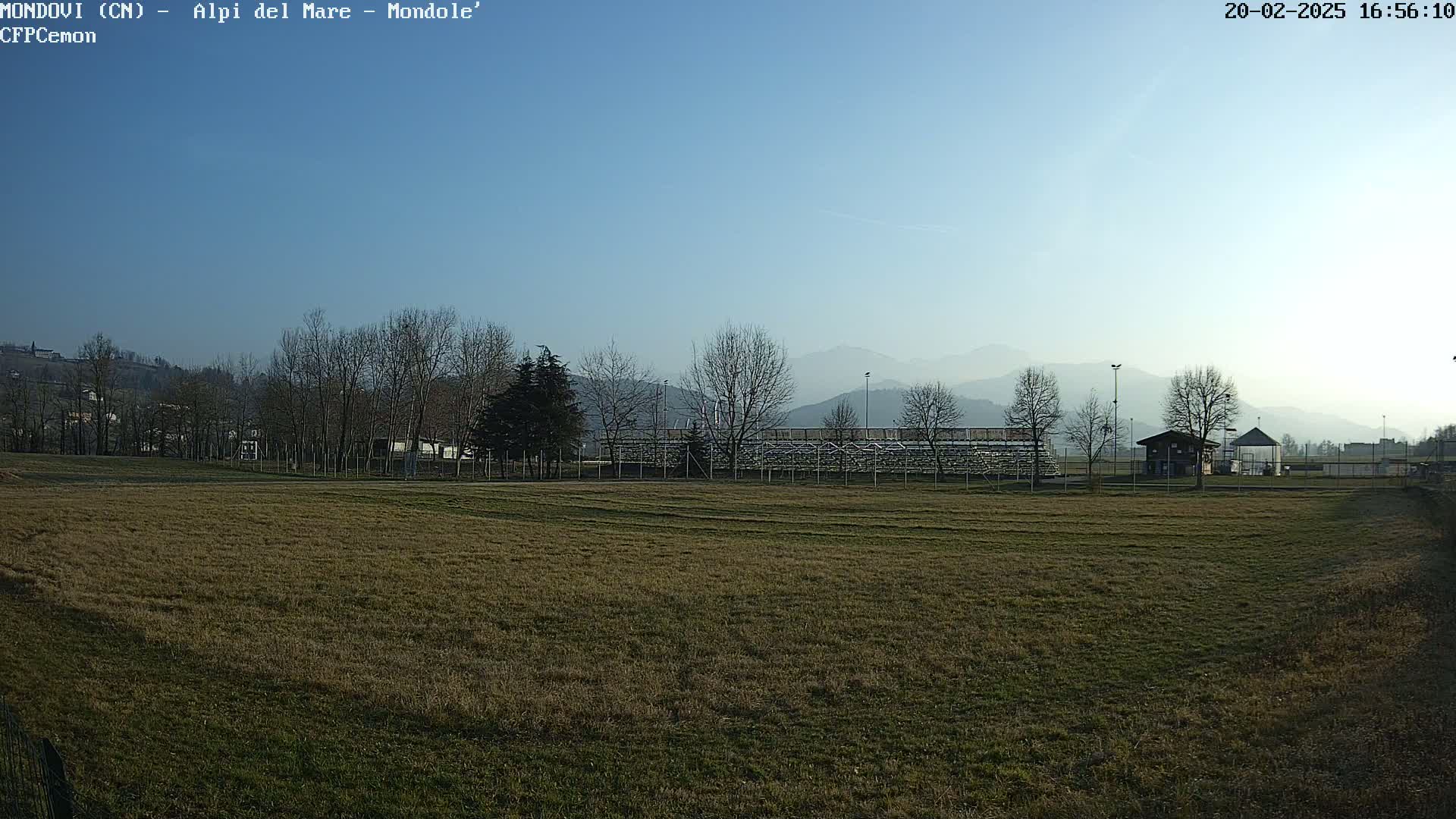 A brown grassy field stretches towards bleachers and a small building, with bare trees and hazy mountains in the background under a clear blue sky.