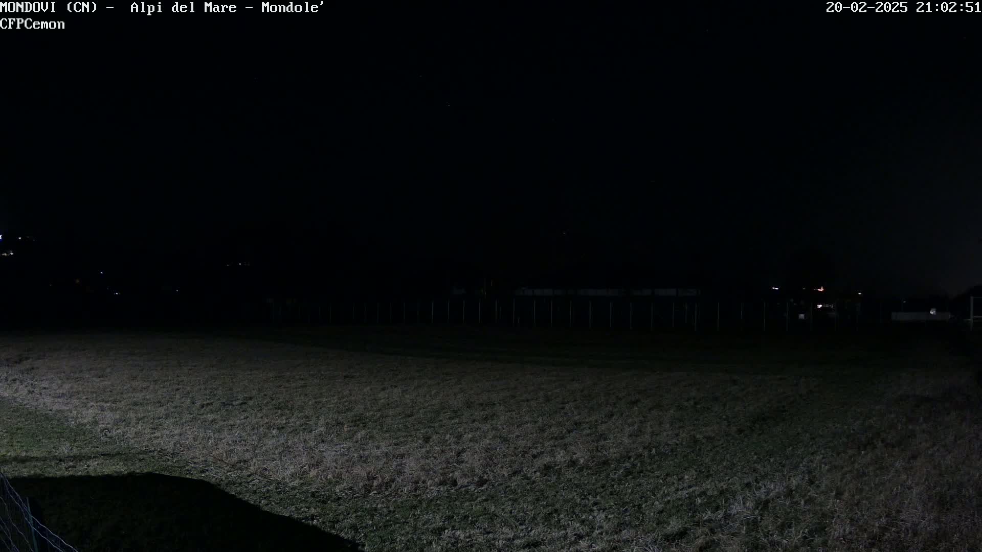 A dark, nighttime view of a grassy field enclosed by a fence, with dimly lit structures and a few faint lights visible in the distance under a clear sky.