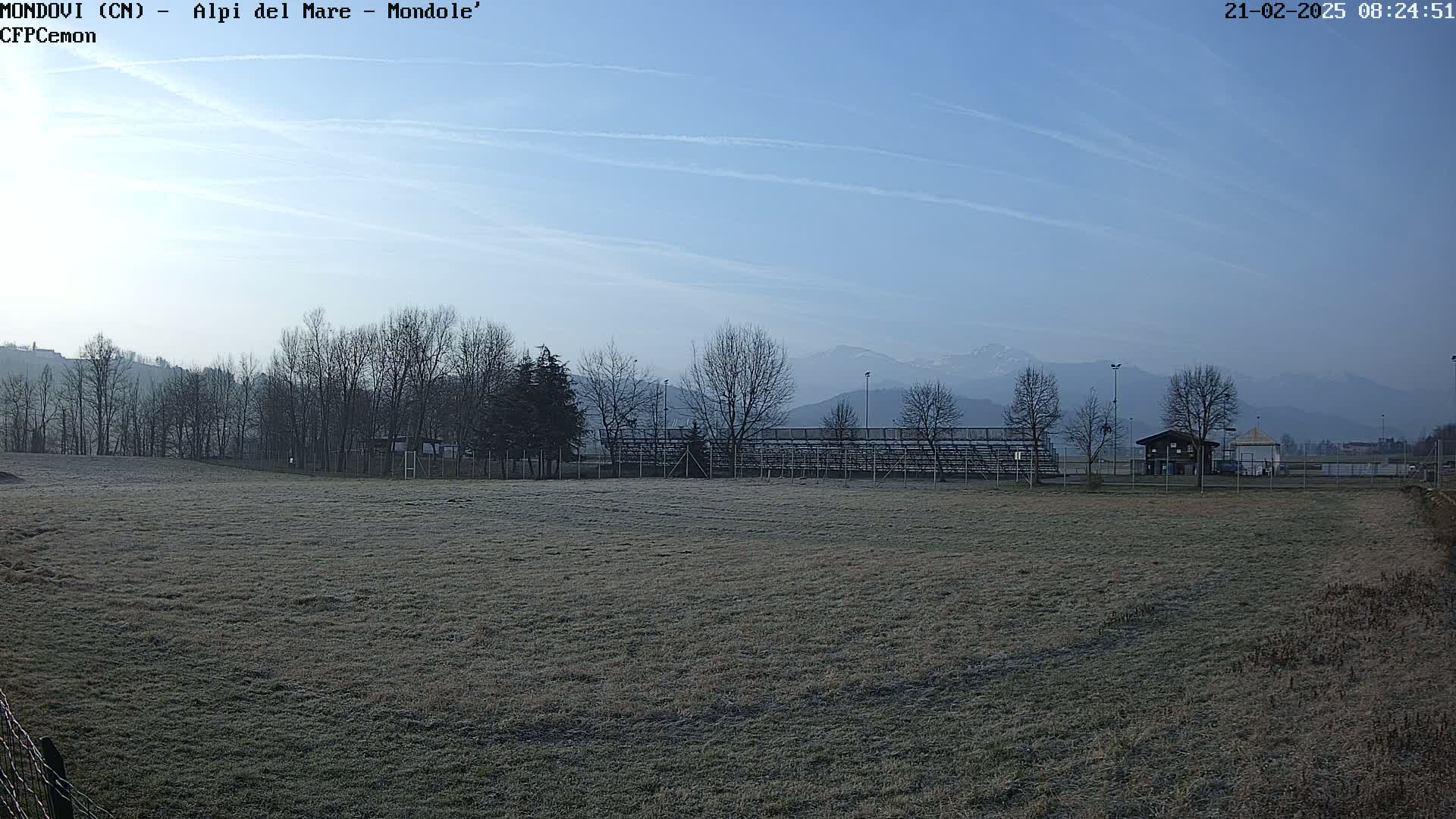 A frost-covered field stretches toward a line of leafless trees and empty bleachers, with snow-capped mountains visible in the distance under a clear, bright sky.