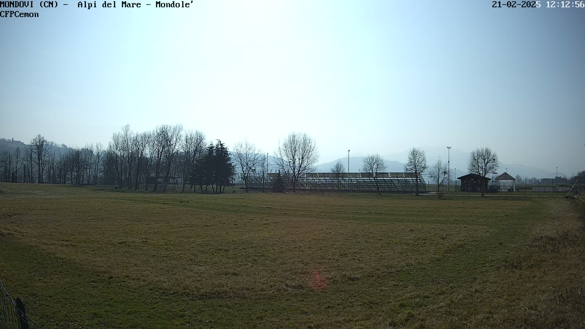A mostly sunny day reveals a brown grassy field with bare trees and a small stadium bleacher structure in the distance, hazy mountains in the background.