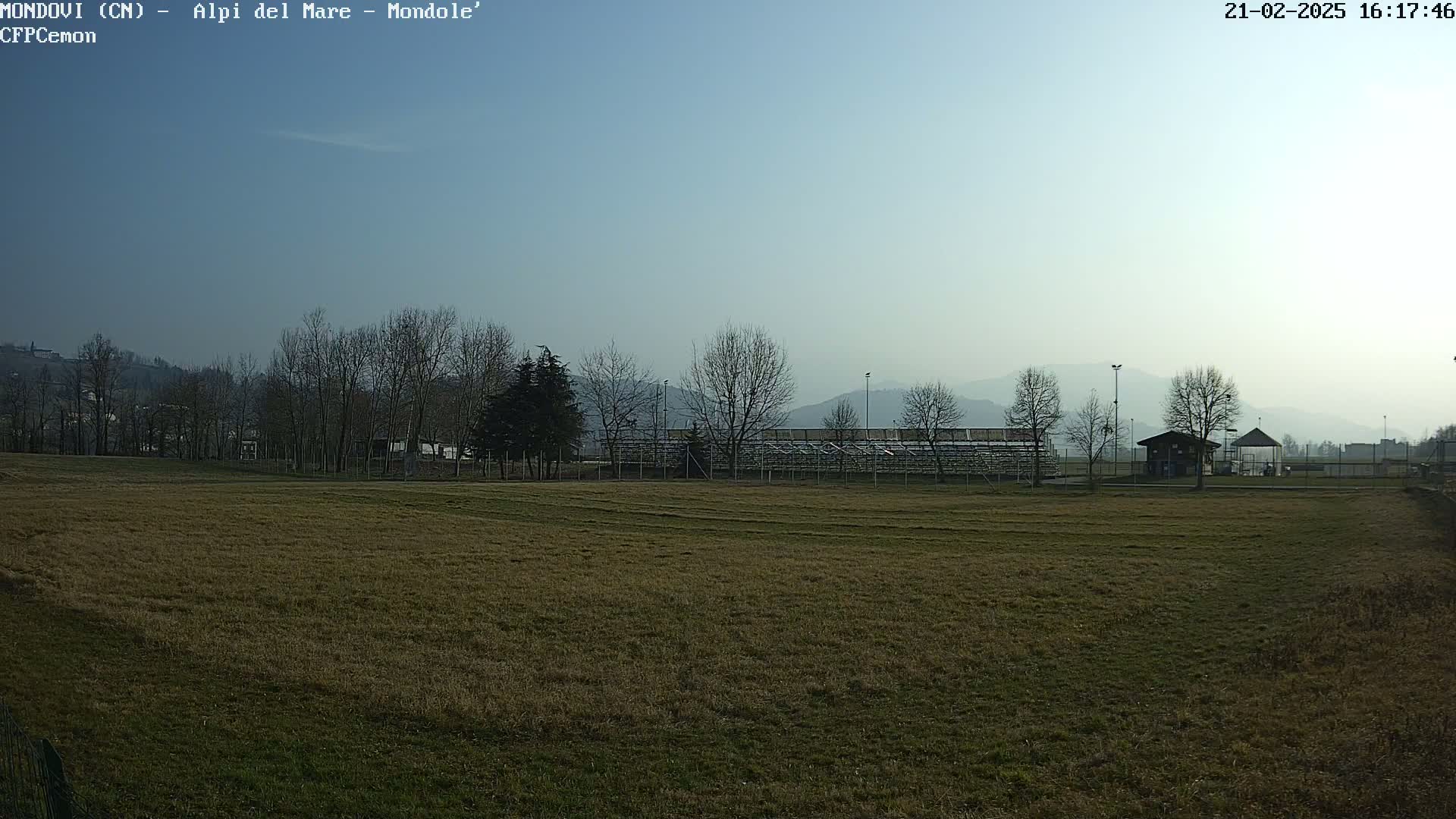 A brown, grassy field stretches out before bleachers and a small building, all situated beneath a clear, pale blue sky and distant, hazy mountains.