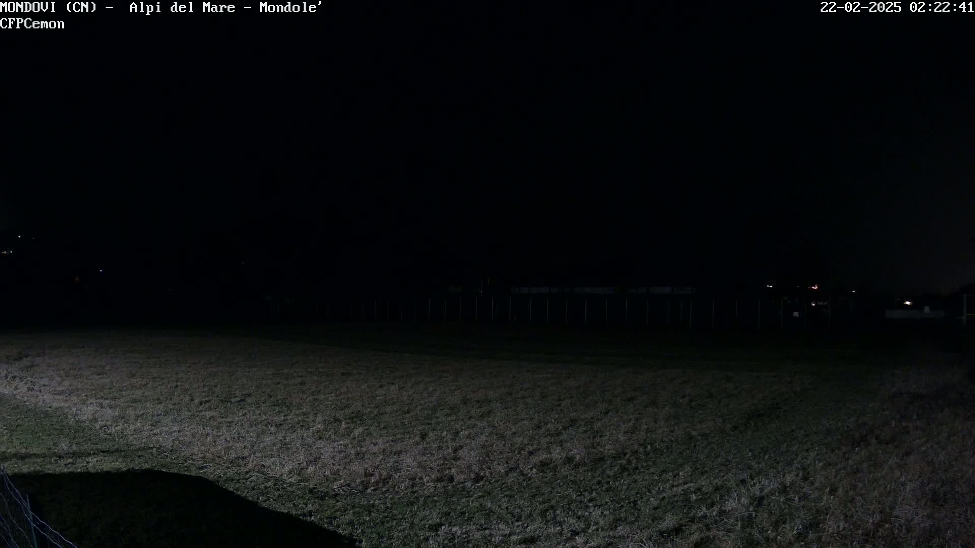 A dark, nighttime view of a grassy field with a fence and distant, faint lights.