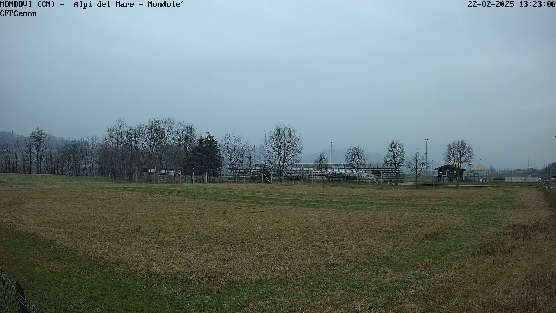 A large, grassy field stretches toward bleachers and a small building under an overcast sky.