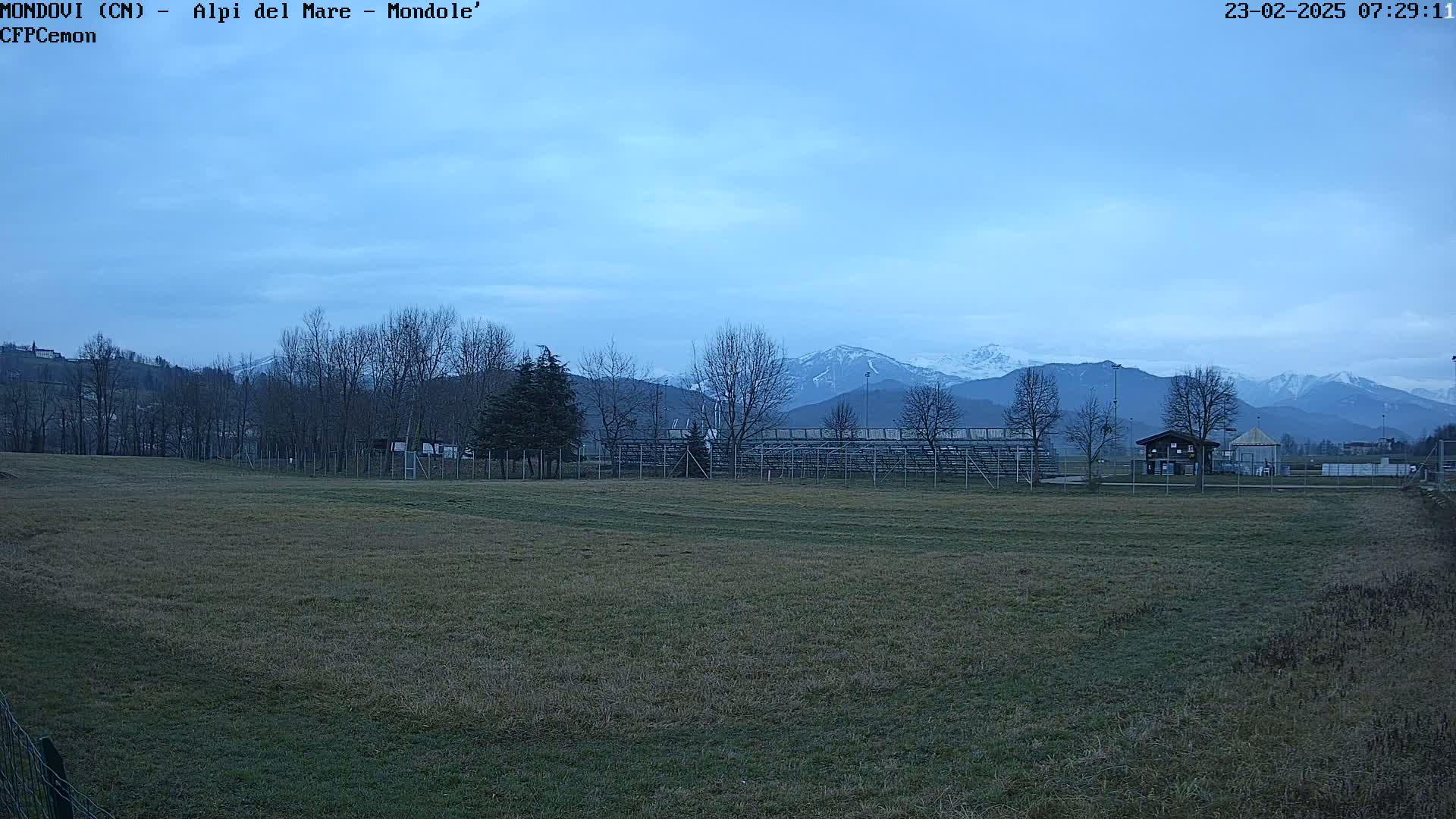 A snow-capped mountain range is visible in the distance behind a grassy field, bleachers, and a few bare trees under a mostly clear, pale blue sky.