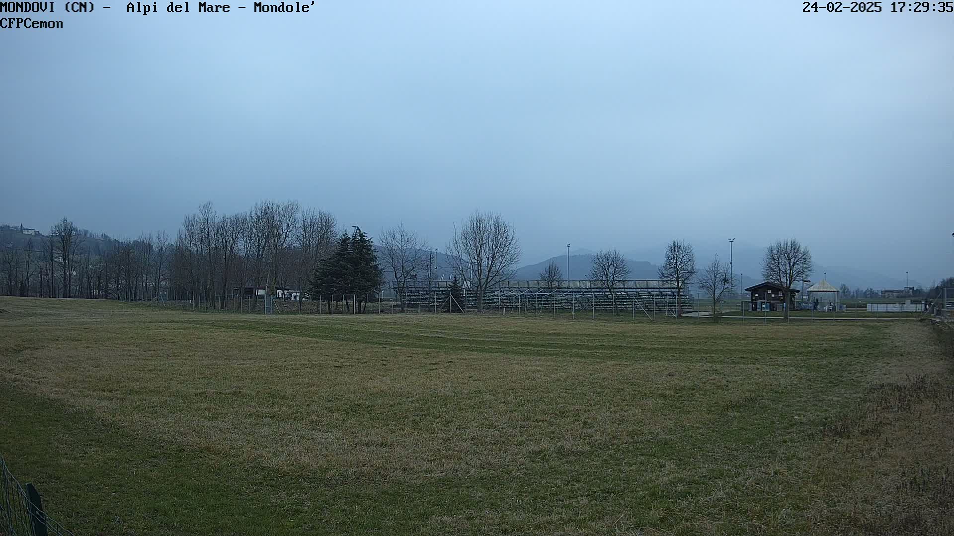 A mostly clear, overcast sky hangs over a grassy field with bleachers and a small building in the background, and bare trees line the horizon.