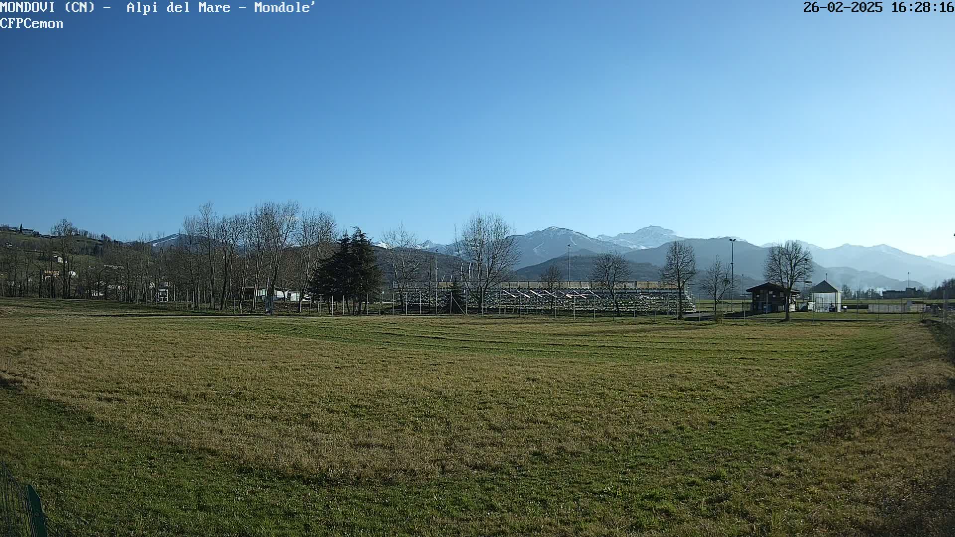 A grassy field stretches out before a small structure and bleachers, all set against a backdrop of snow-capped mountains under a clear blue sky.