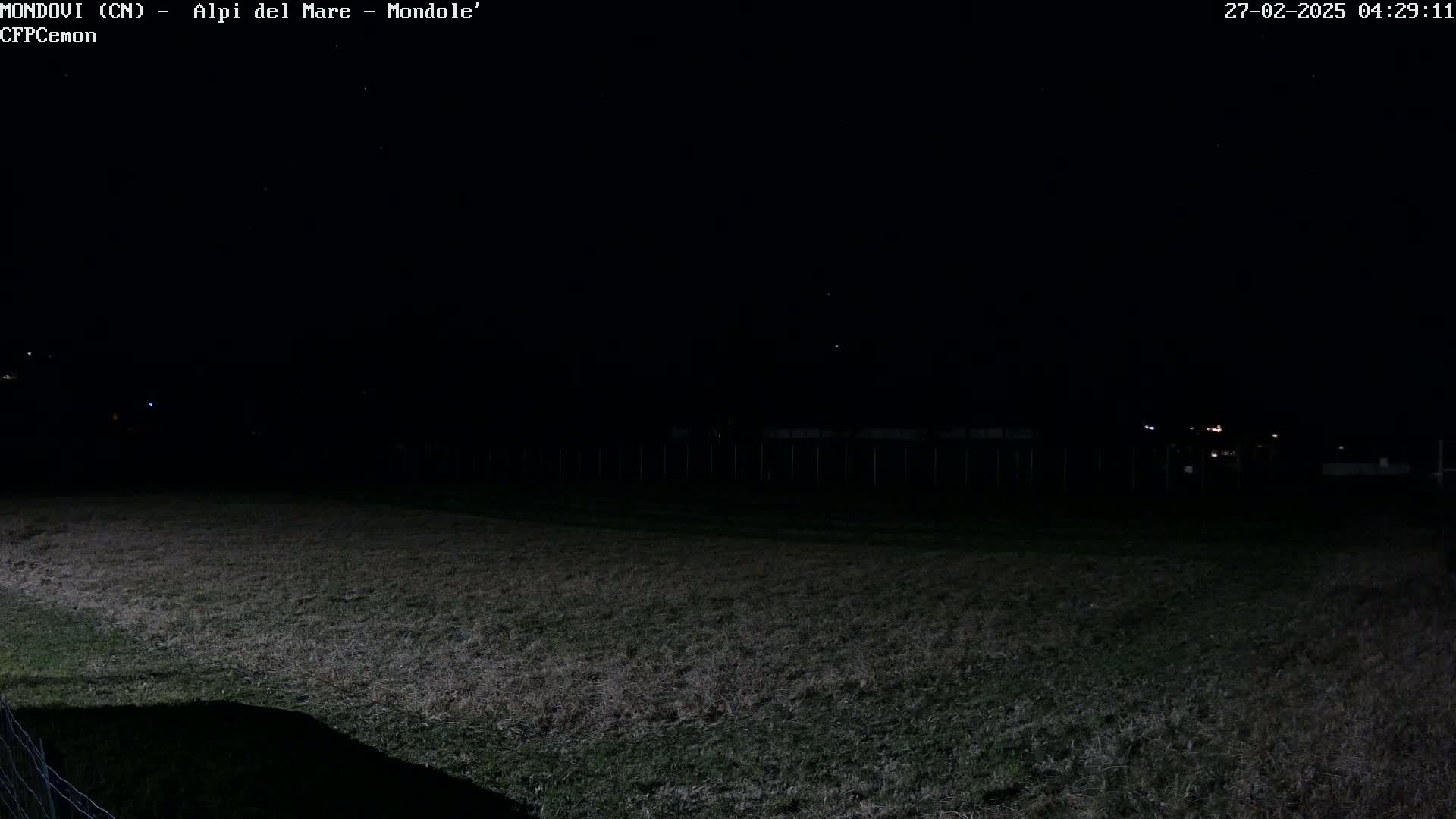 A dark, nighttime view of a grassy field extending to a fence under a clear, starlit sky.