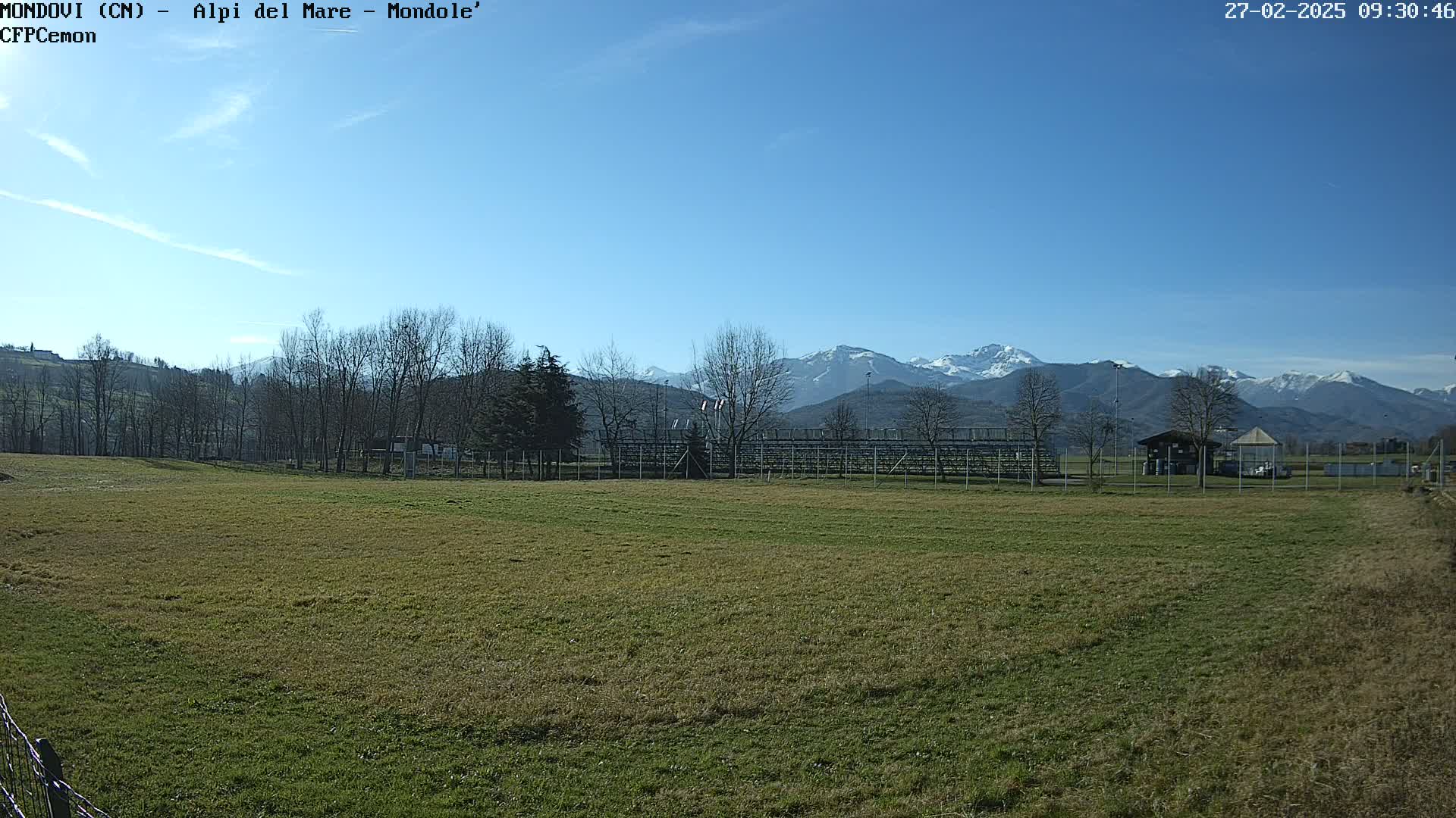 A grassy field with bleachers and a small building sits before a line of leafless trees and snow-capped mountains under a clear blue sky.