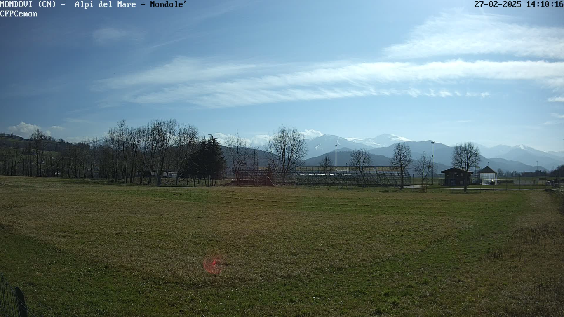 A grassy field with a few trees and a small building, overlooks a snow-capped mountain range under a mostly sunny sky.