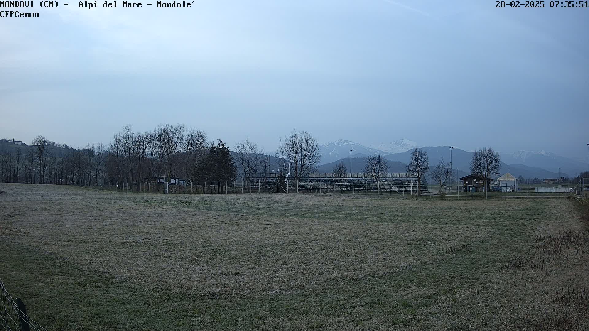A field with frost-covered grass, bleachers, and a few buildings, overlooks snow-capped mountains under a pale, overcast sky.
