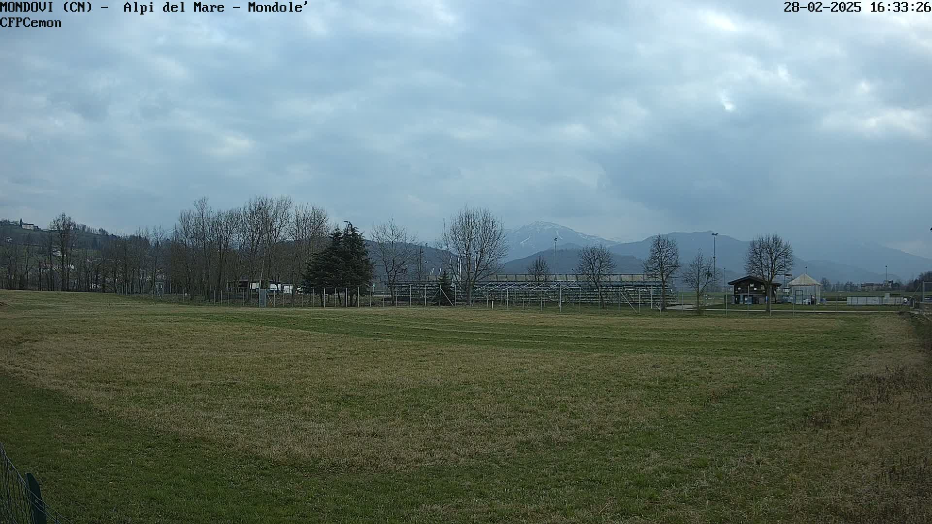 A grassy field stretches toward bleachers and a small building, with leafless trees and snow-capped mountains visible in the distance under a cloudy sky.