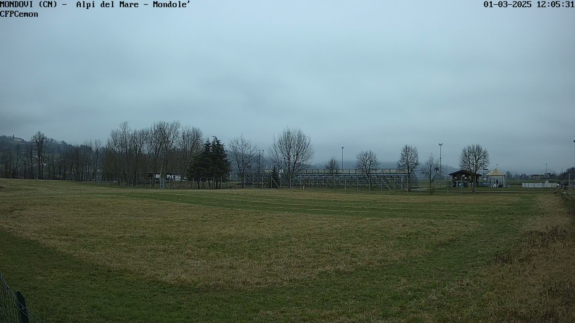 A grassy field, under an overcast sky, stretches towards bleachers and bare trees in the mid-ground, with a small structure and hills in the distance.