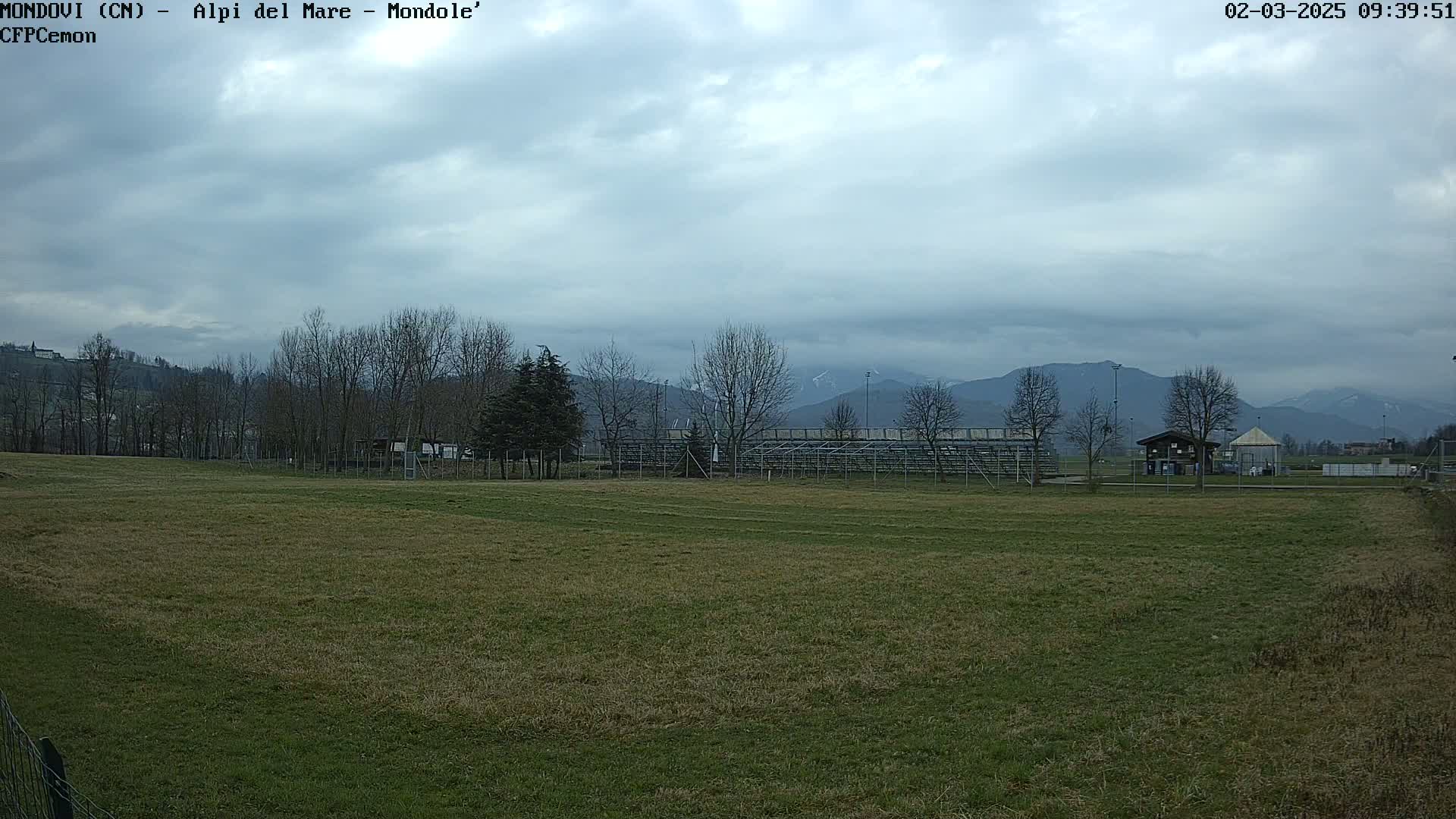 A grassy field with bleachers and a small building sits in the foreground, under a cloudy sky, with mountains in the background.