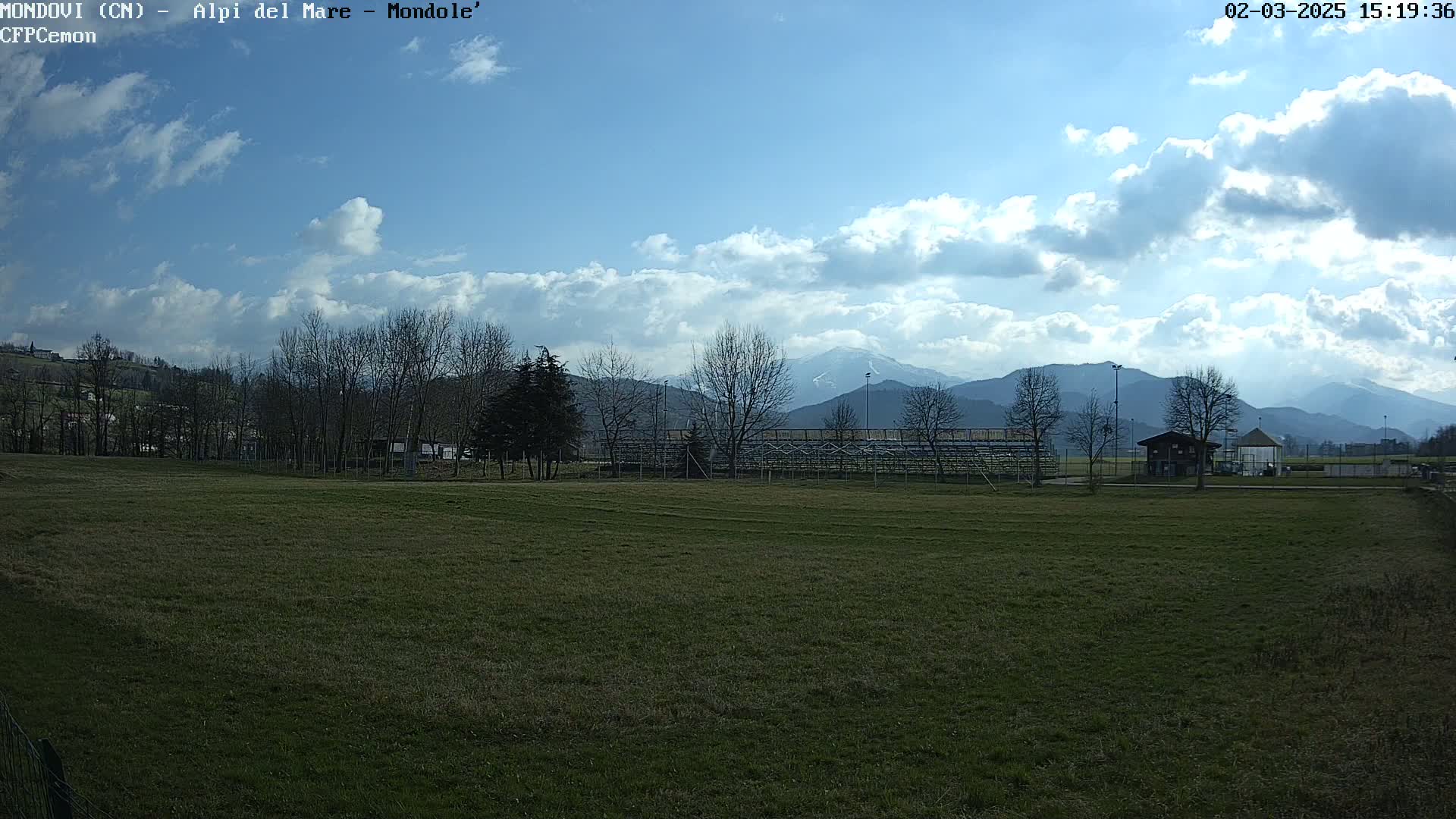 A grassy field sits before bleachers and a small building, with bare trees and snow-capped mountains in the background under a partly cloudy sky.
