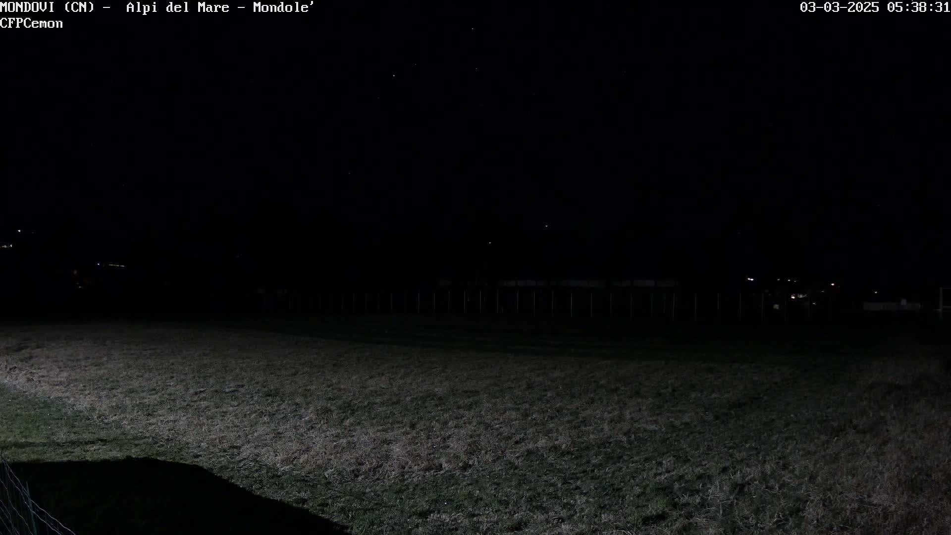 A dark, nighttime view of a grassy field with a fence and distant lights under a clear, starlit sky.