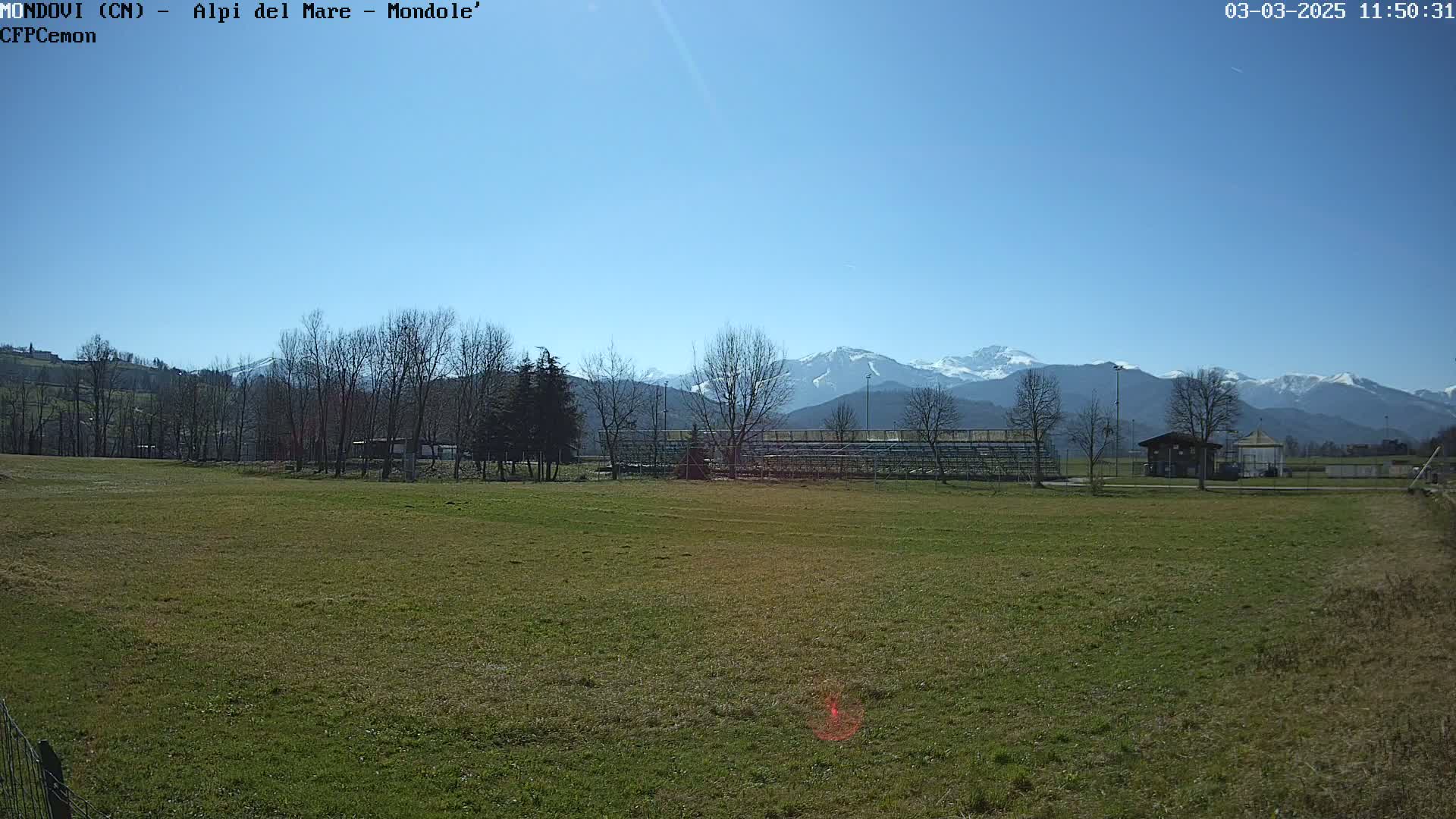 A grassy field with bleachers and a small building in the midground, under a clear blue sky, with snow-capped mountains in the background.