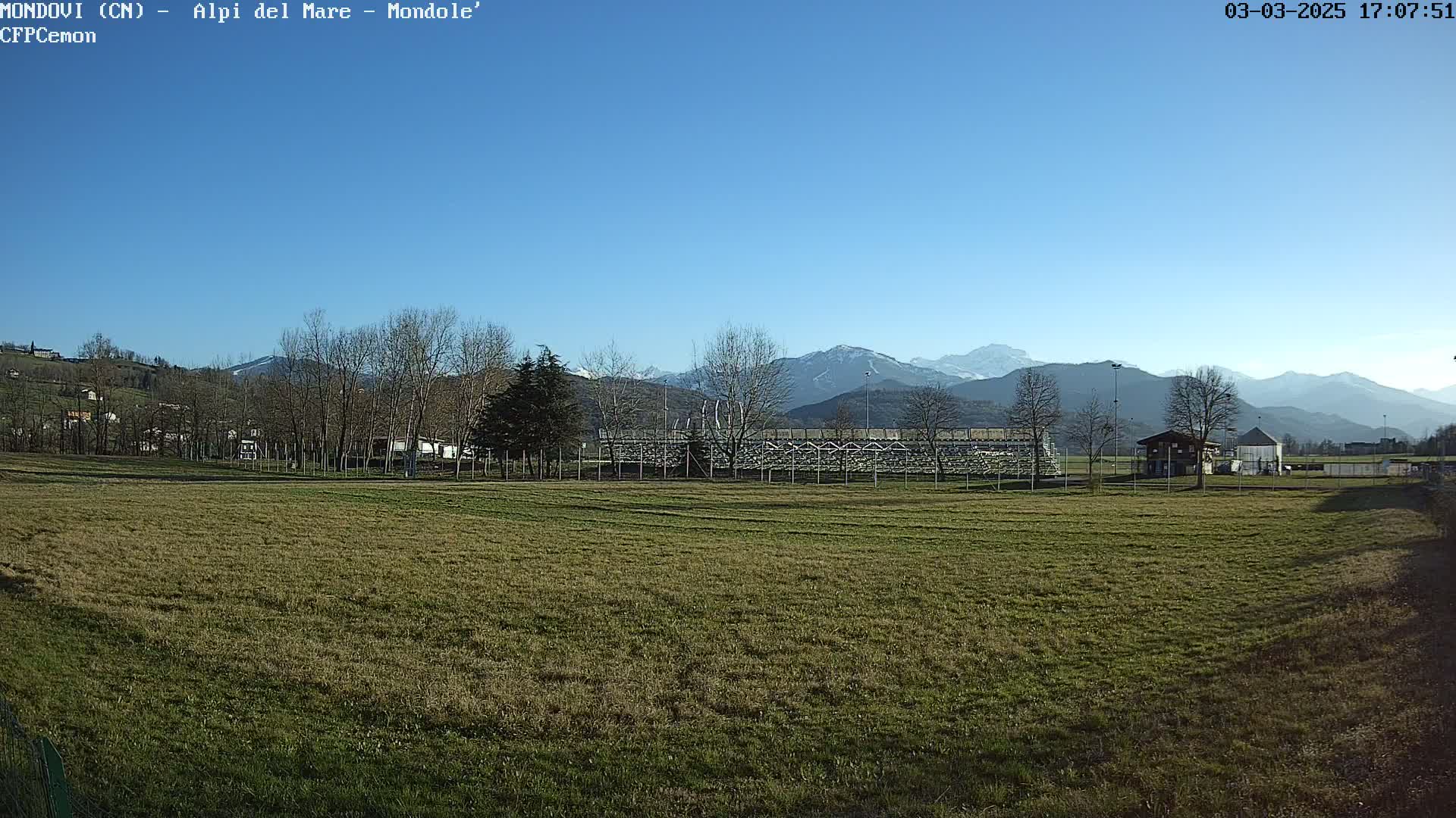 A grassy field stretches towards a fenced-in area with bleachers, beyond which are leafless trees and snow-capped mountains under a clear blue sky.