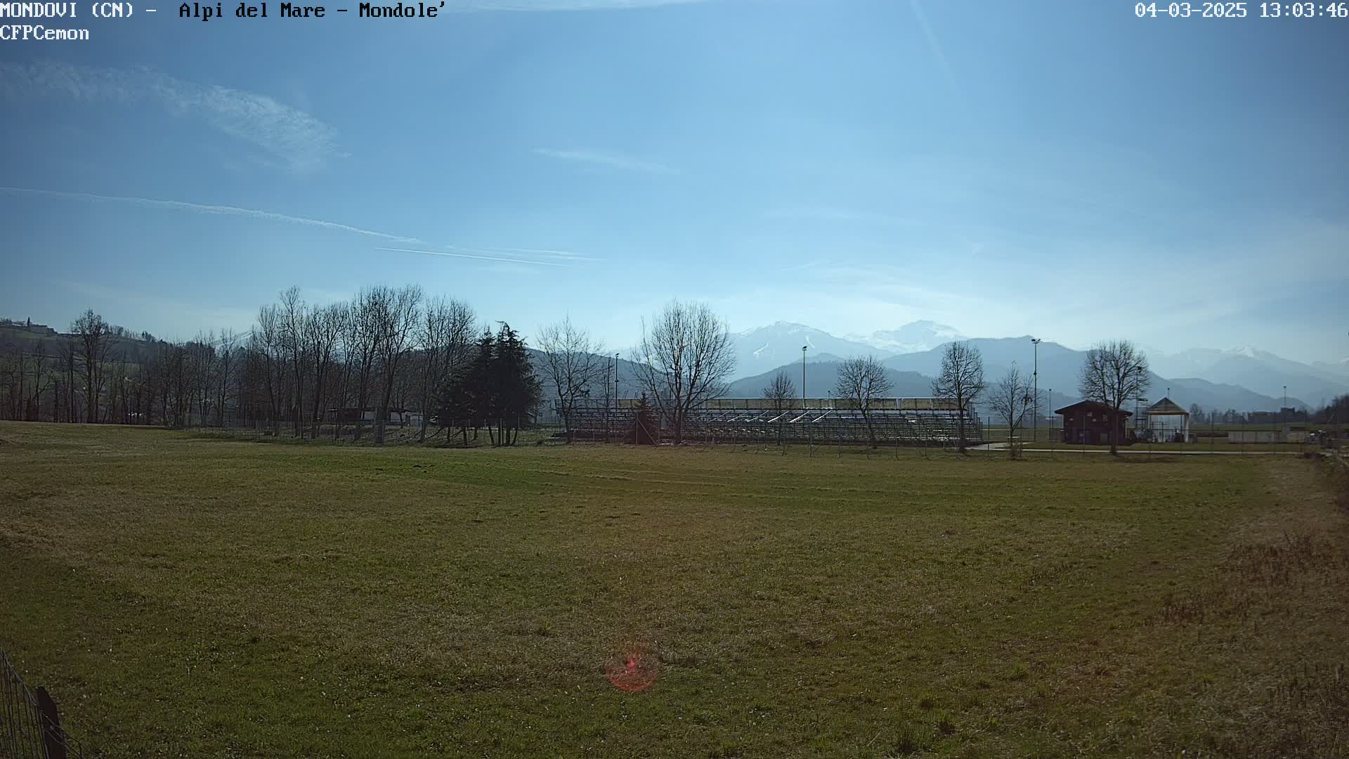 A grassy field with bleachers and a small building sits in the foreground, overlooking a line of leafless trees and snow-capped mountains under a clear, sunny sky.