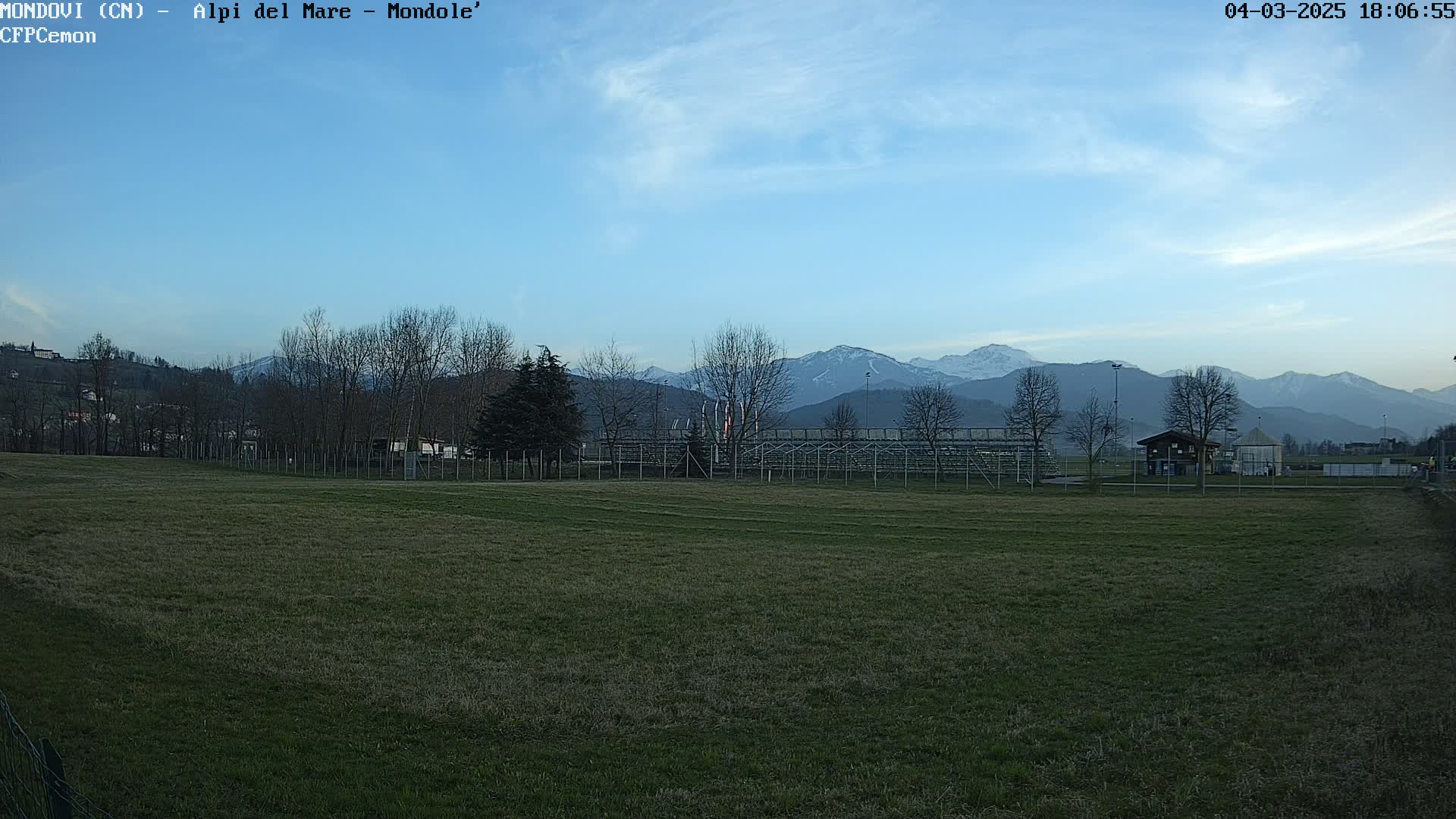 A grassy field with bleachers and a small building sits in the foreground, under a mostly clear sky, with snow-capped mountains in the background.