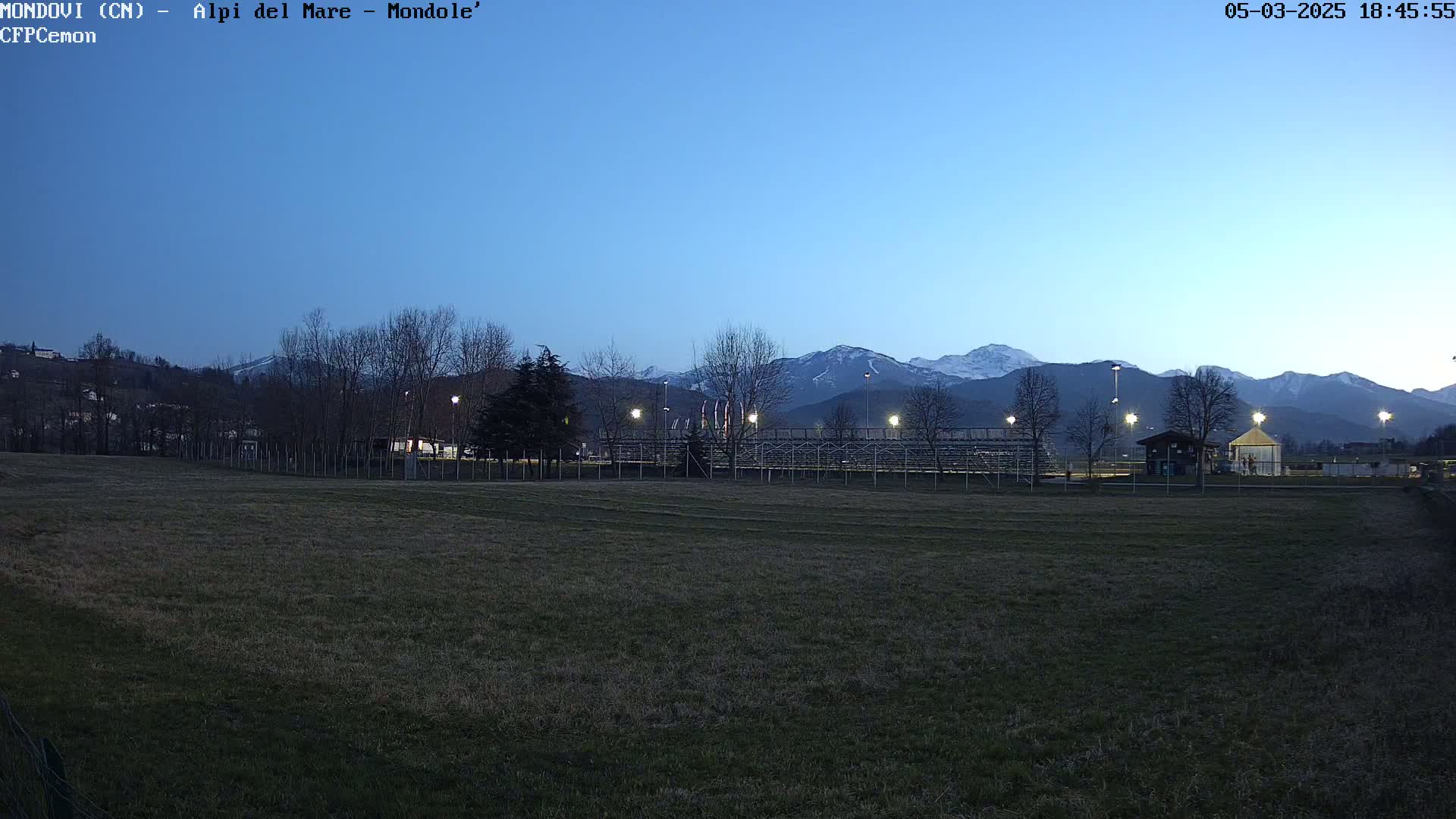 A mostly empty field with bleachers and a small building sits in front of snow-capped mountains under a twilight sky.