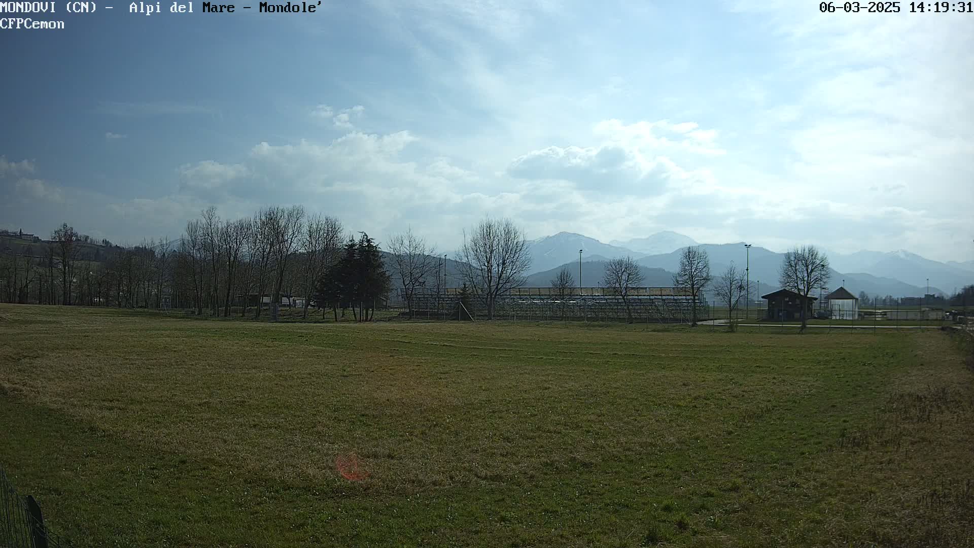 A grassy field with leafless trees and a small building sits before a distant mountain range under a partly cloudy sky.