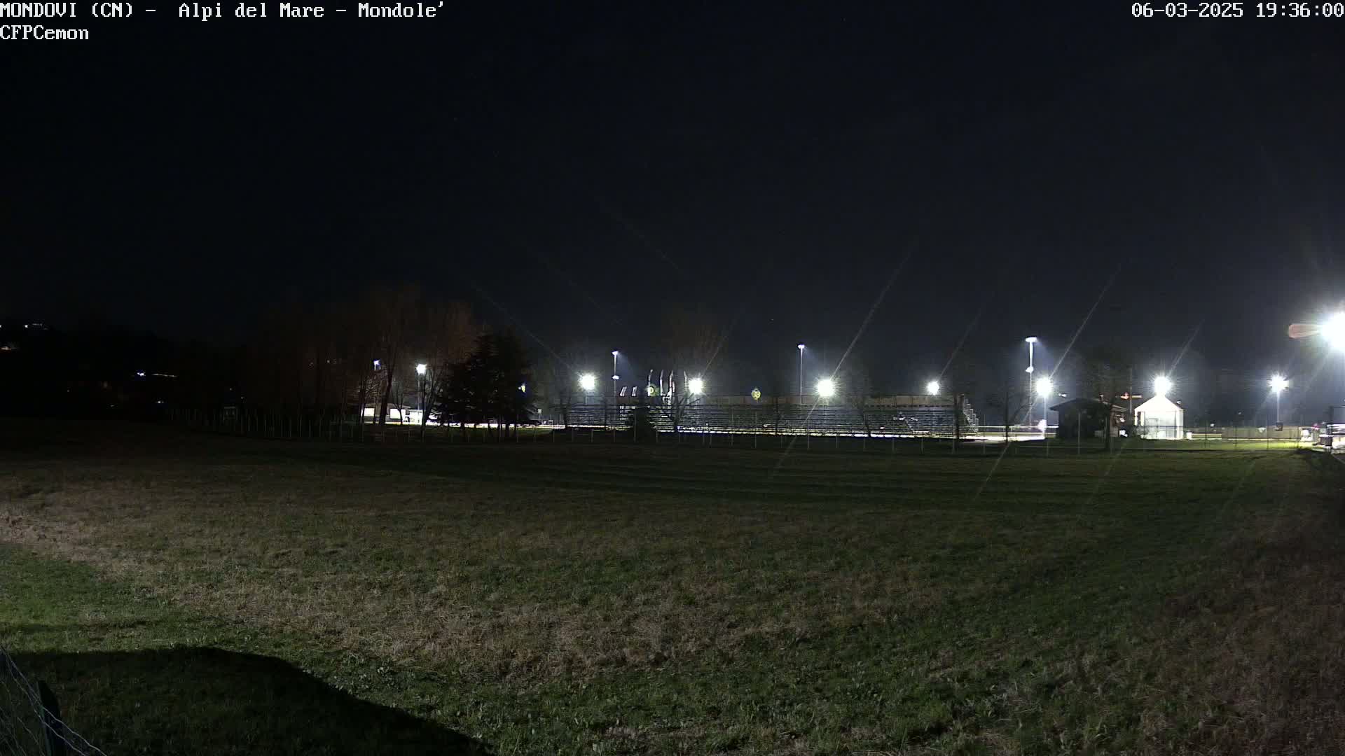 A nighttime, outdoor view of a grassy field with a lit-up sports stadium in the background.