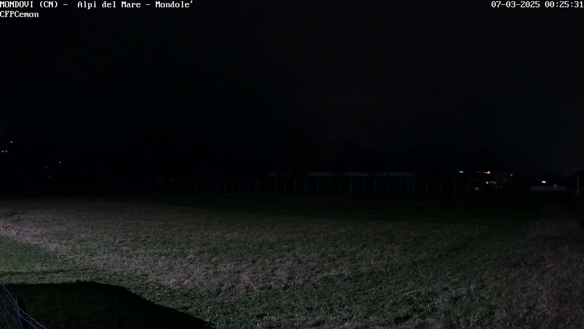 A dark, nighttime view of a grassy field enclosed by a fence, with dimly lit structures and distant lights visible in the background.