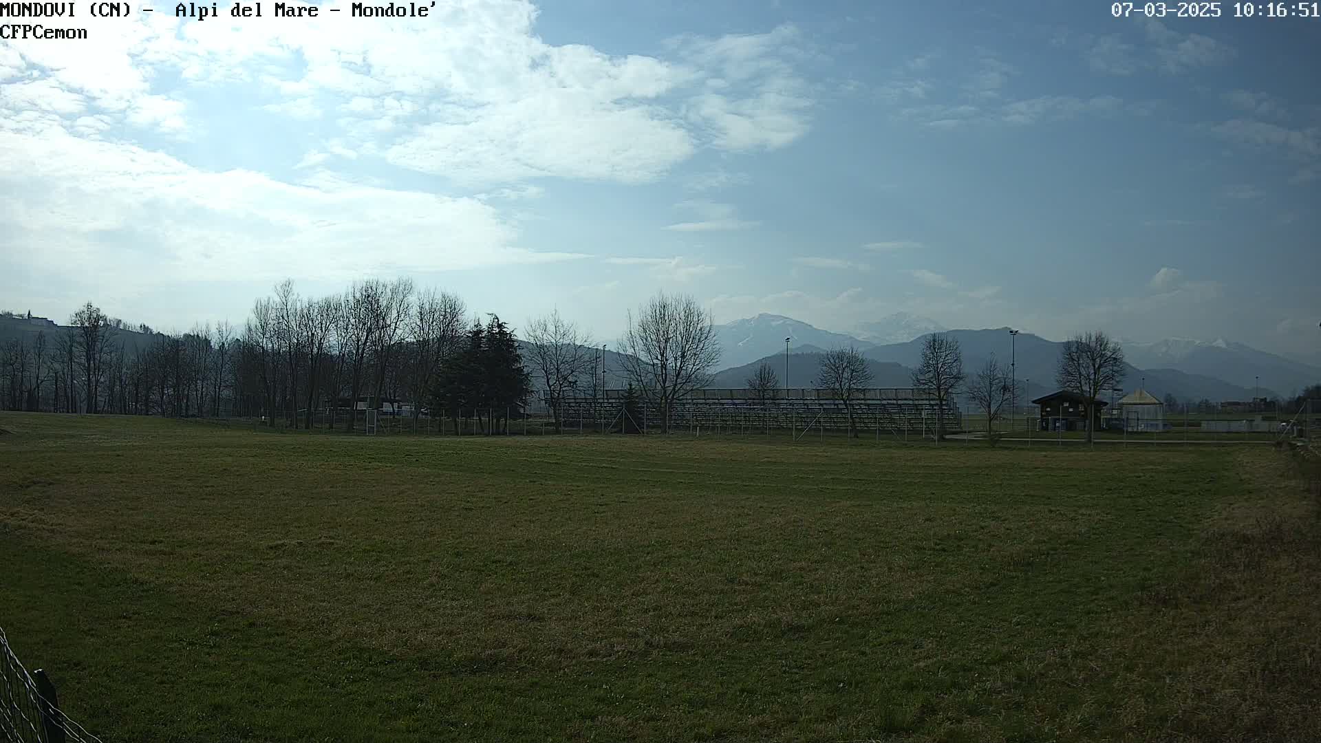 A grassy field with bleachers and a small building sits before a line of bare trees and snow-capped mountains under a partly cloudy sky.