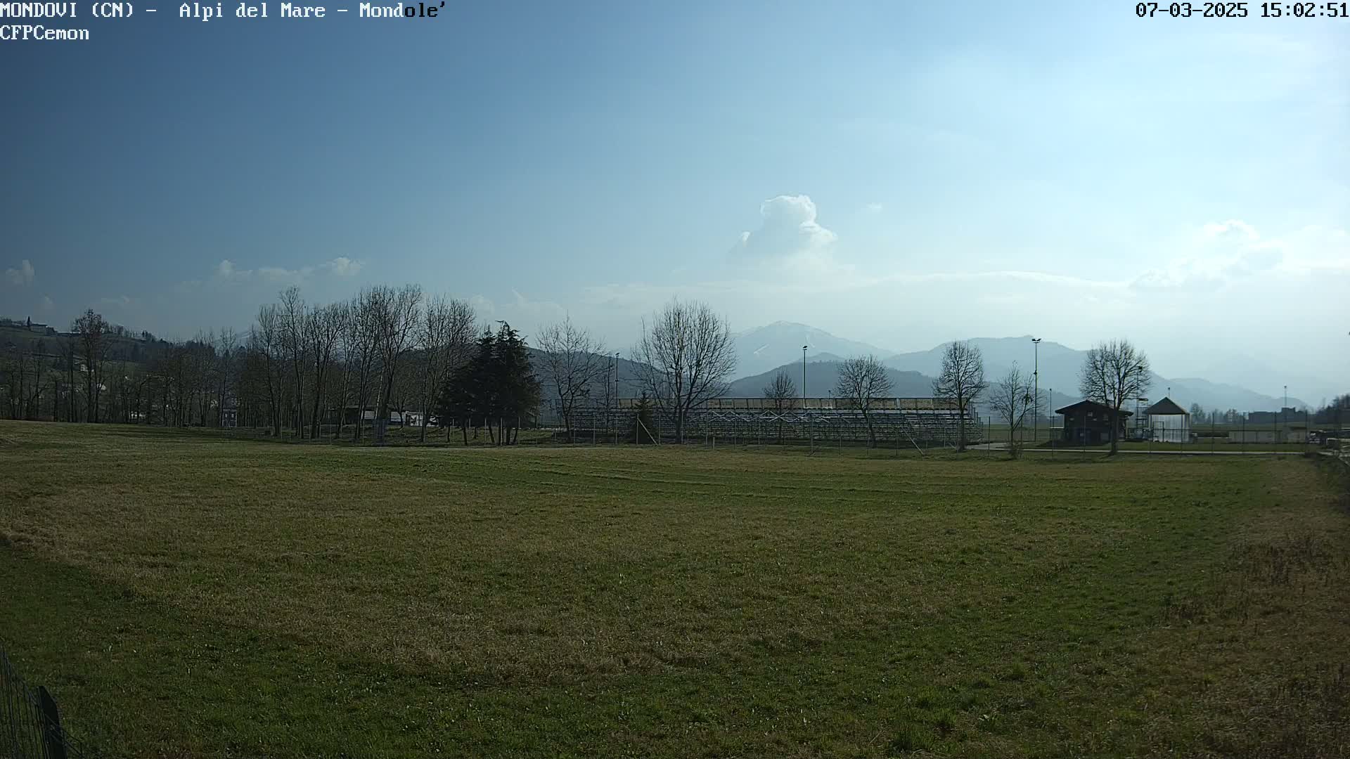 A mostly sunny day reveals a grassy field, leafless trees, a small building, and bleachers, all set against a backdrop of distant mountains.