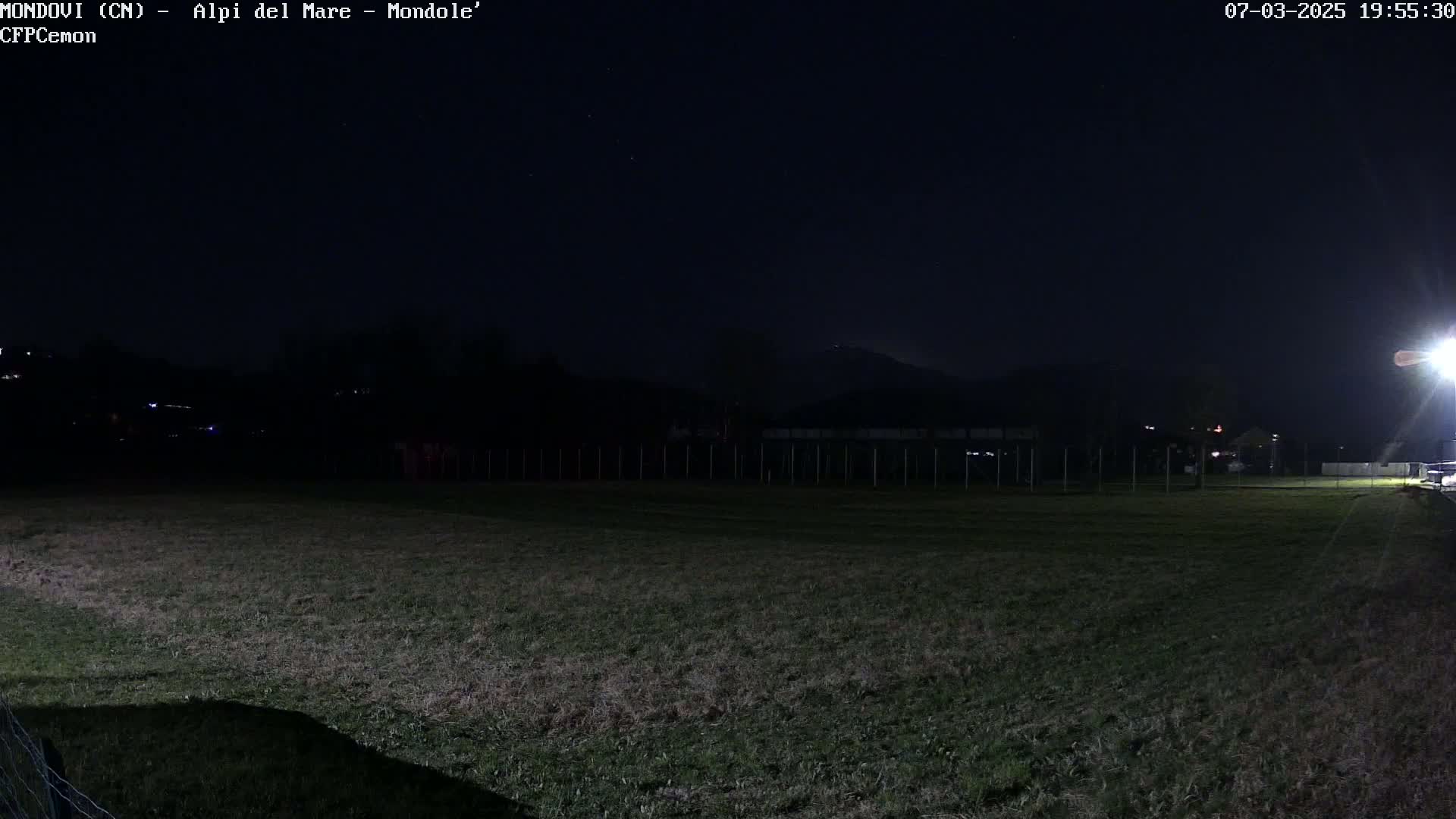 A dark, nighttime scene shows a grassy field with a fence and distant hills under a clear, starry sky.
