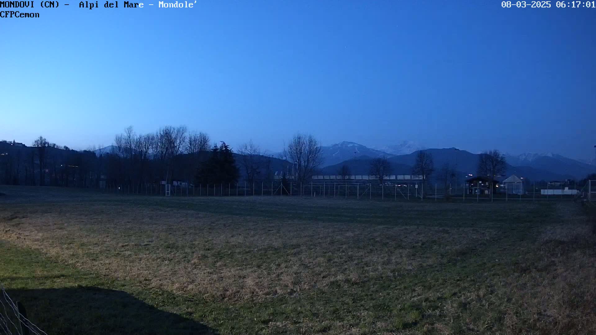 A field with leafless trees and a fence in the foreground, overlooking a distant mountain range under a twilight sky.