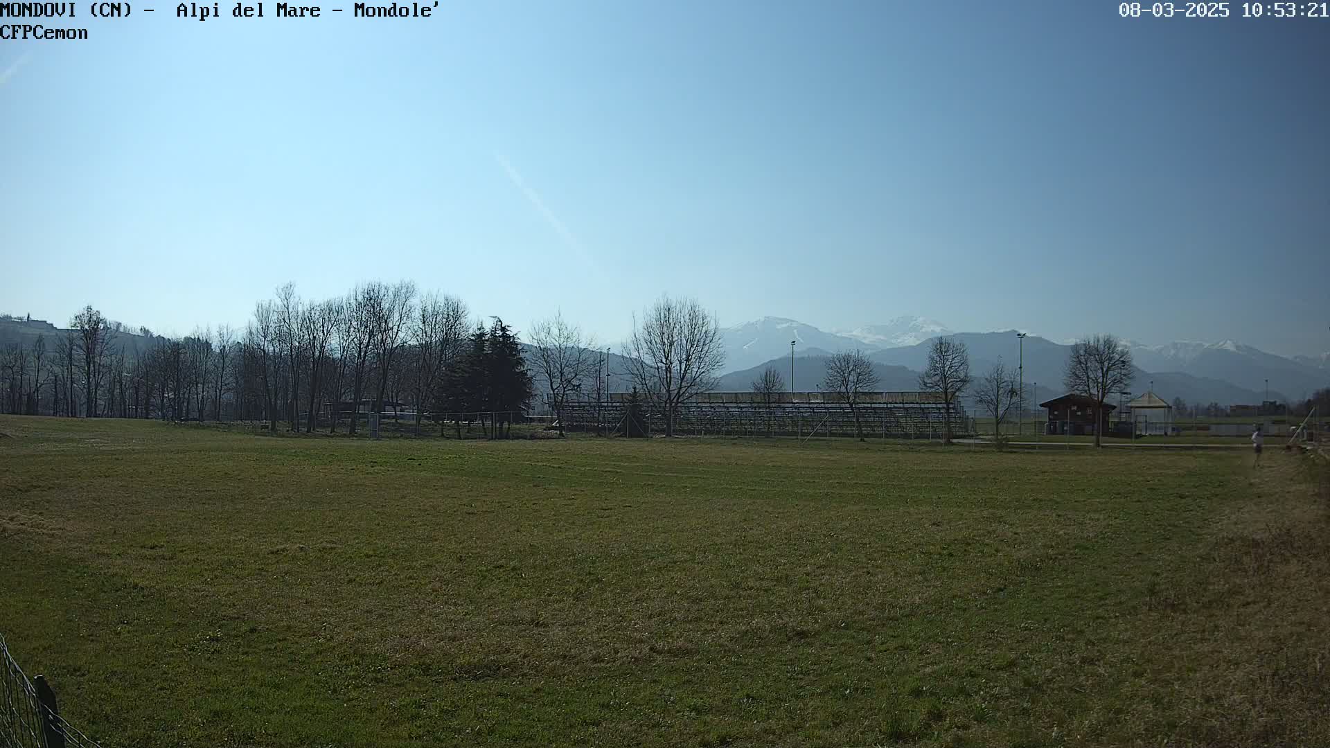 A grassy field stretches toward bleachers and a small building, beyond which bare trees stand before snow-capped mountains under a clear blue sky.