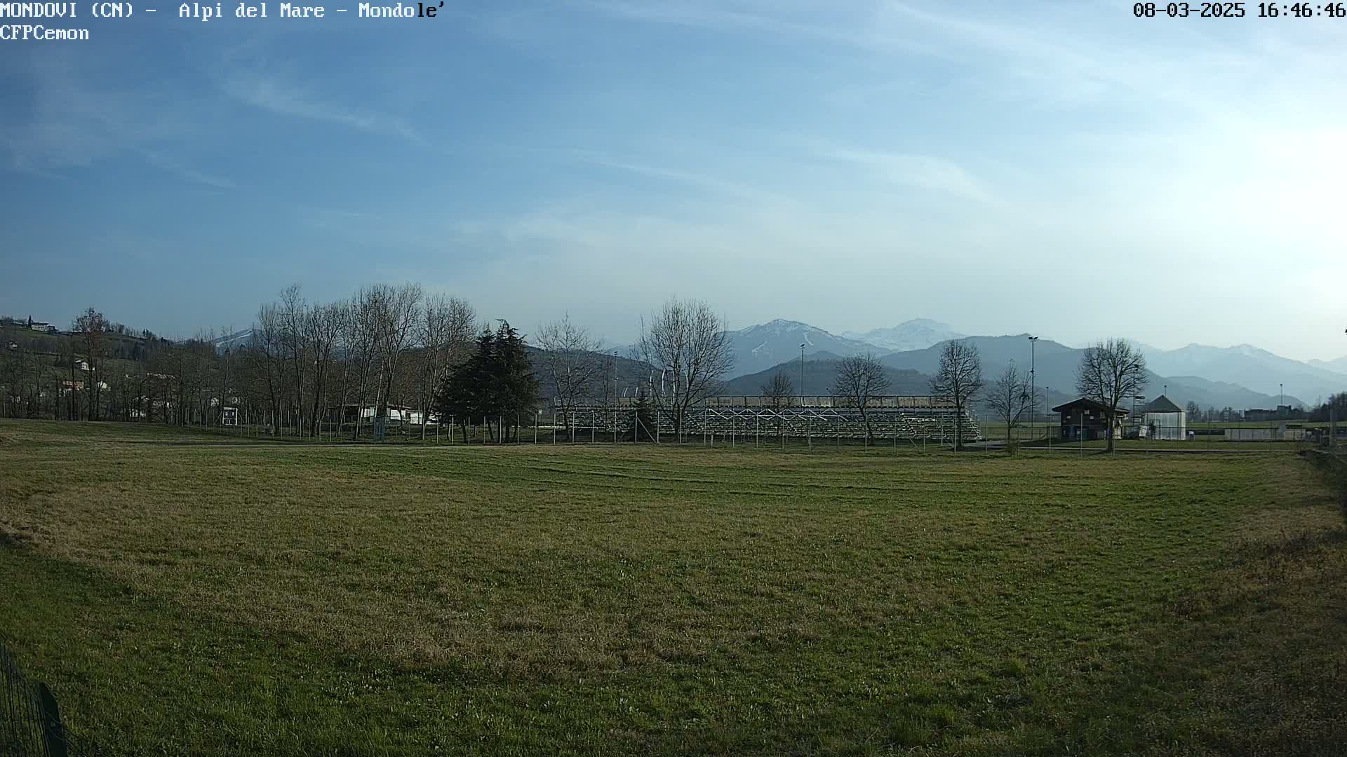 A grassy field with a metal bleacher structure and small buildings is in the foreground, while leafless trees and snow-capped mountains are visible in the background under a mostly clear, bright sky.