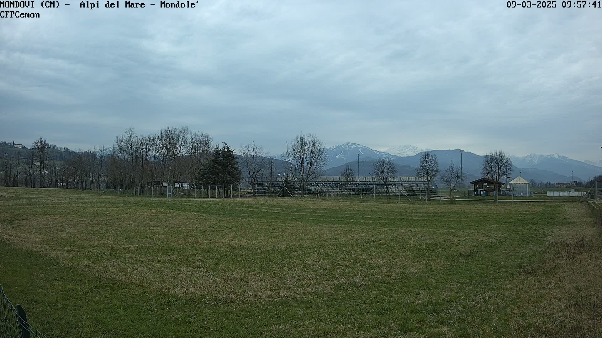 A grassy field with bleachers and a small building sits before snow-capped mountains under a cloudy sky.