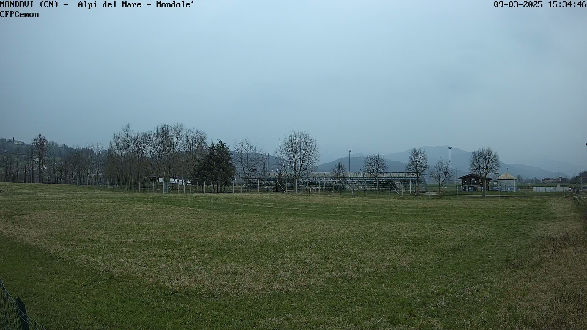 A grassy field with bleachers and a small building is visible in the mid-ground, backed by bare trees and hazy mountains under an overcast sky.